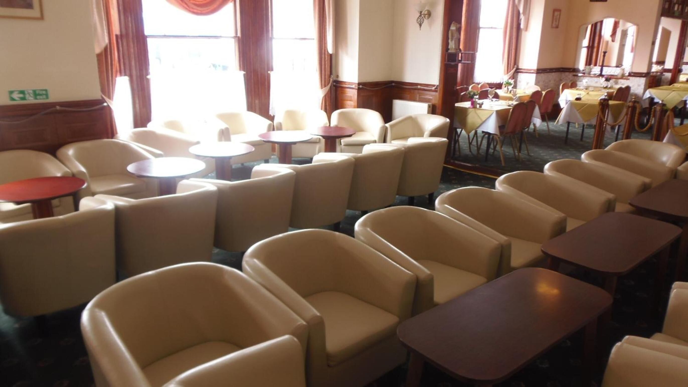 Un hall d’hôtel avec des rangées de fauteuils beiges et de petites tables rondes, un espace repas avec des nappes jaunes et des chaises rouges, et une grande fenêtre avec des rideaux.
