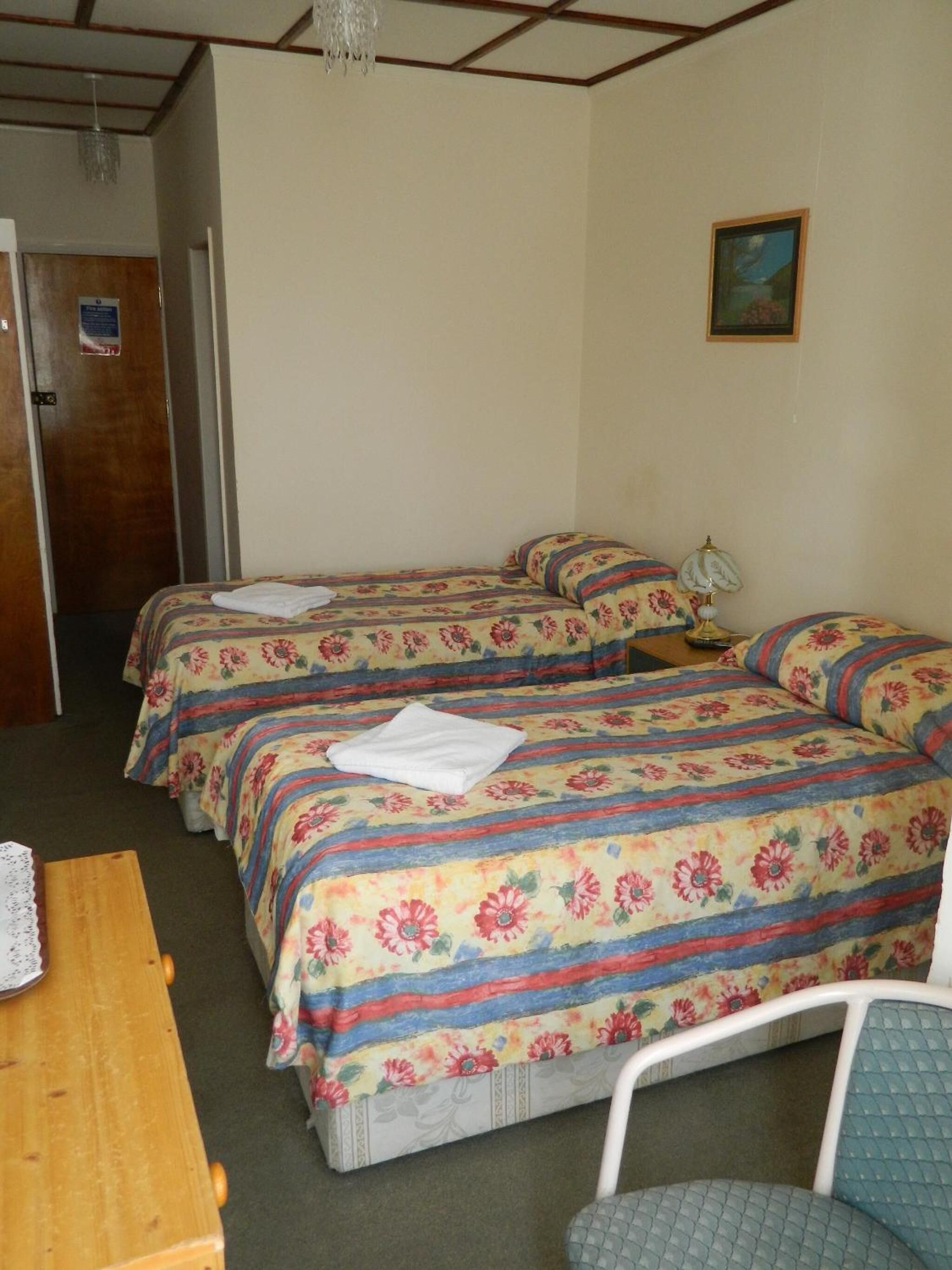 A room with two beds, a wooden table, a chair, a lamp, and a painting on the wall.