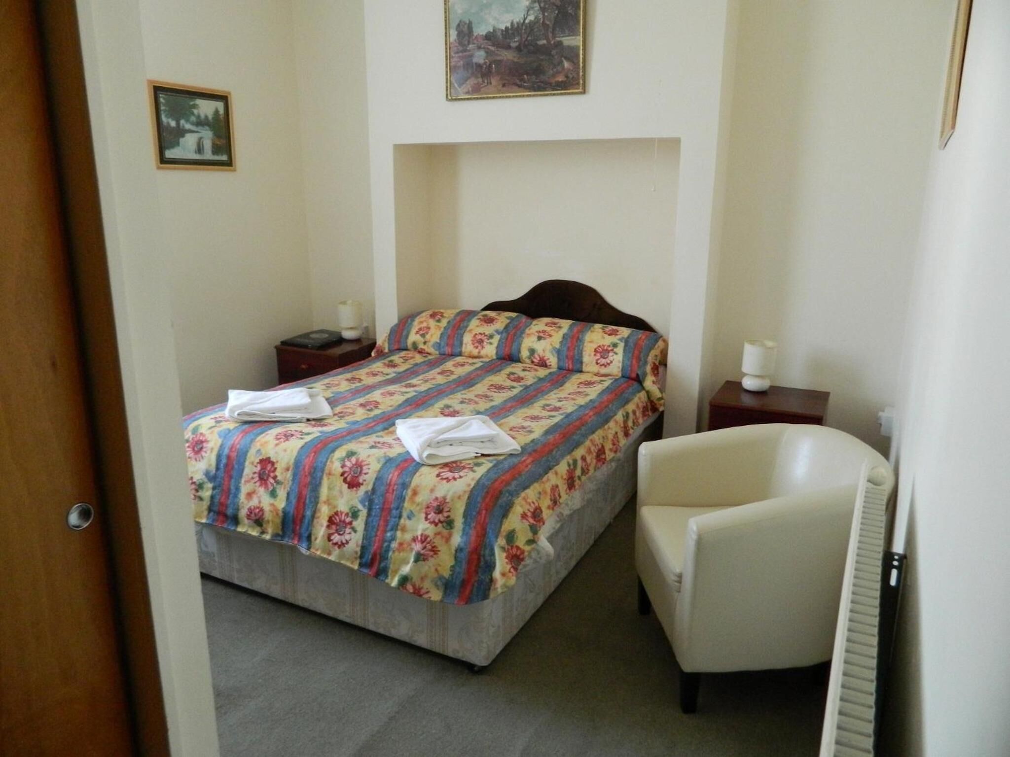 A small, neatly arranged bedroom with a bed, a chair, a nightstand, and a picture on the wall.