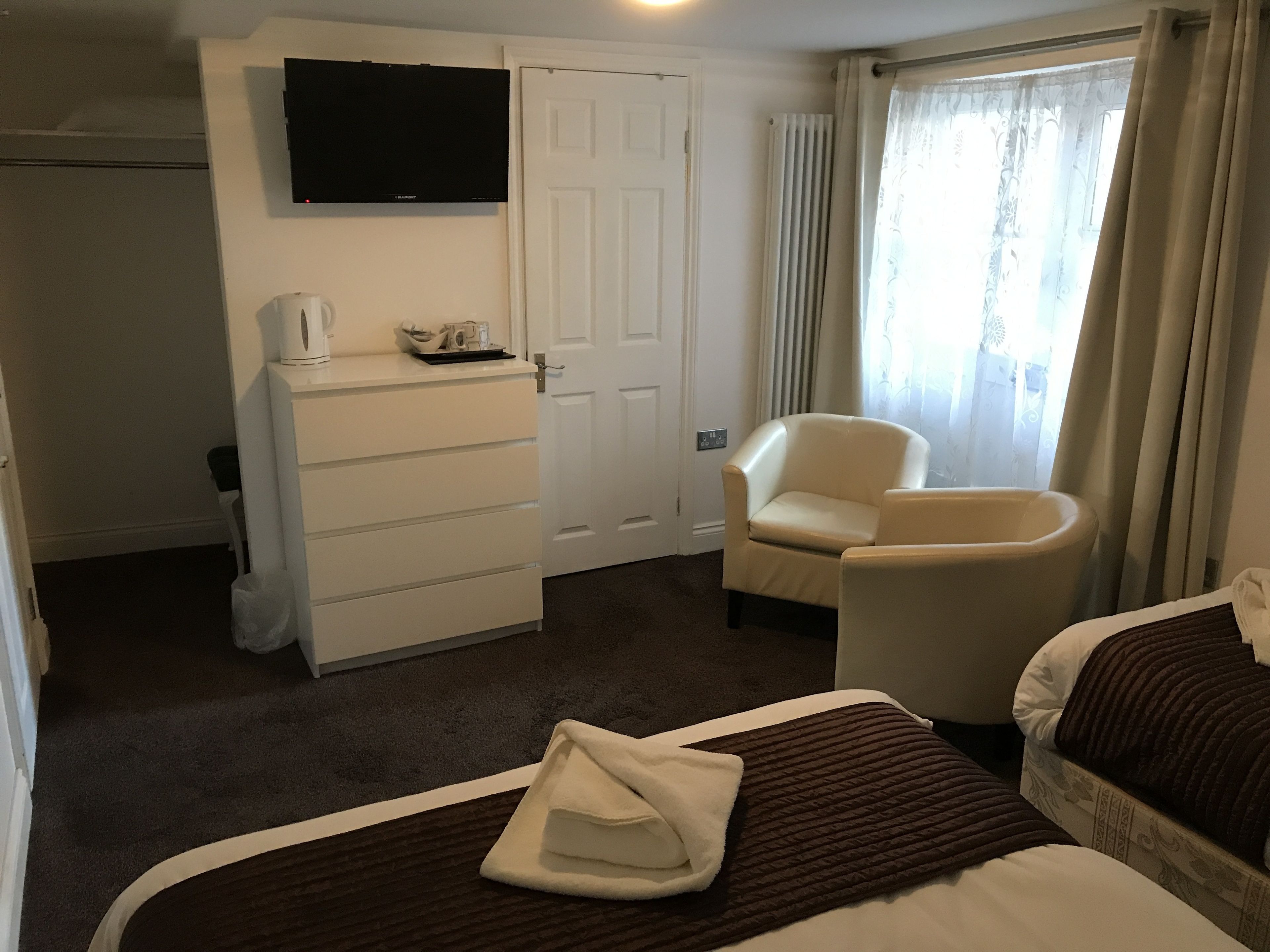A hotel room with a bed, a chair, a desk with a flat-screen TV, a wardrobe, and a window with curtains.