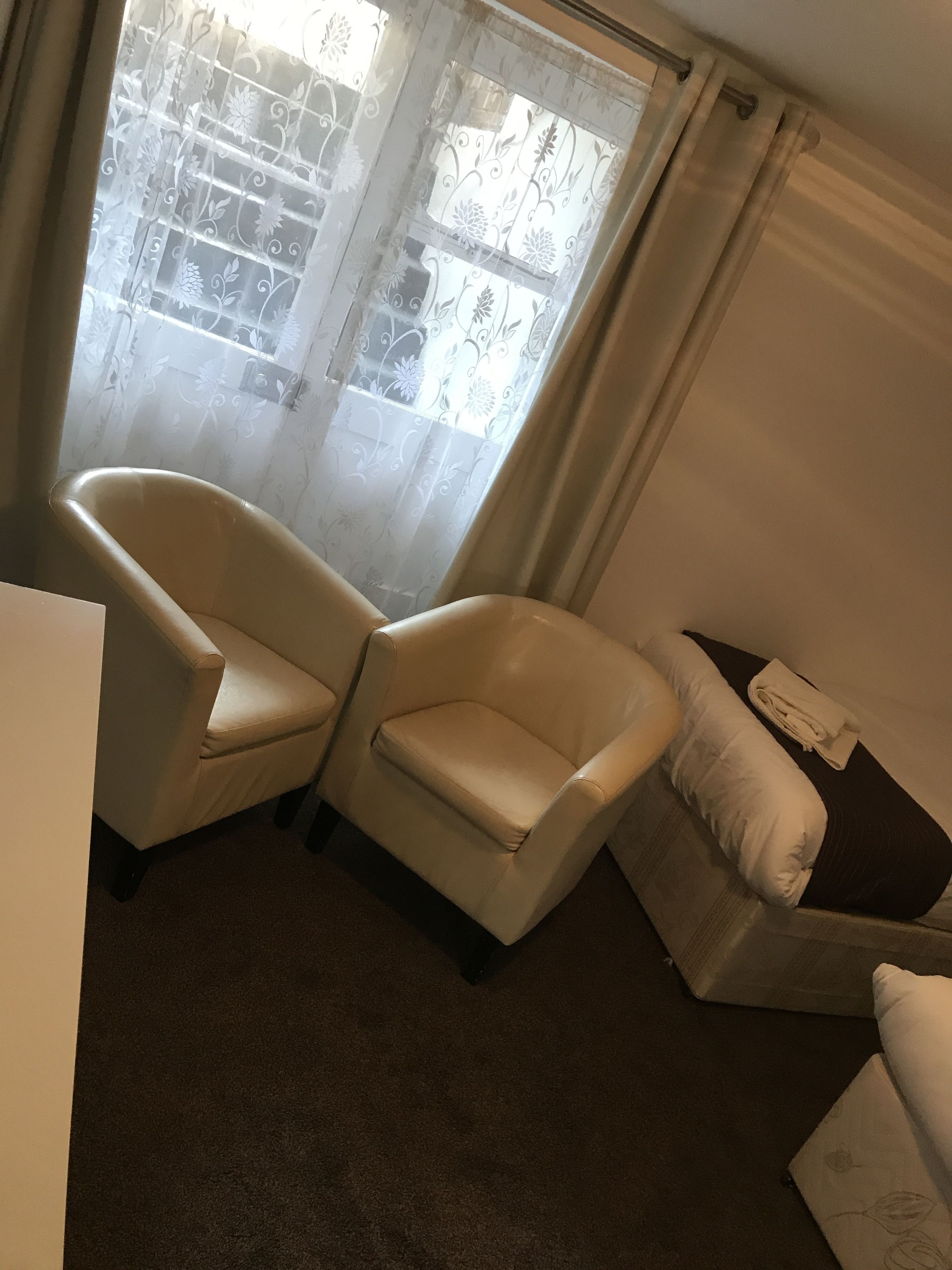 A room with two beige armchairs, a small table, and a bed with a white pillow.
