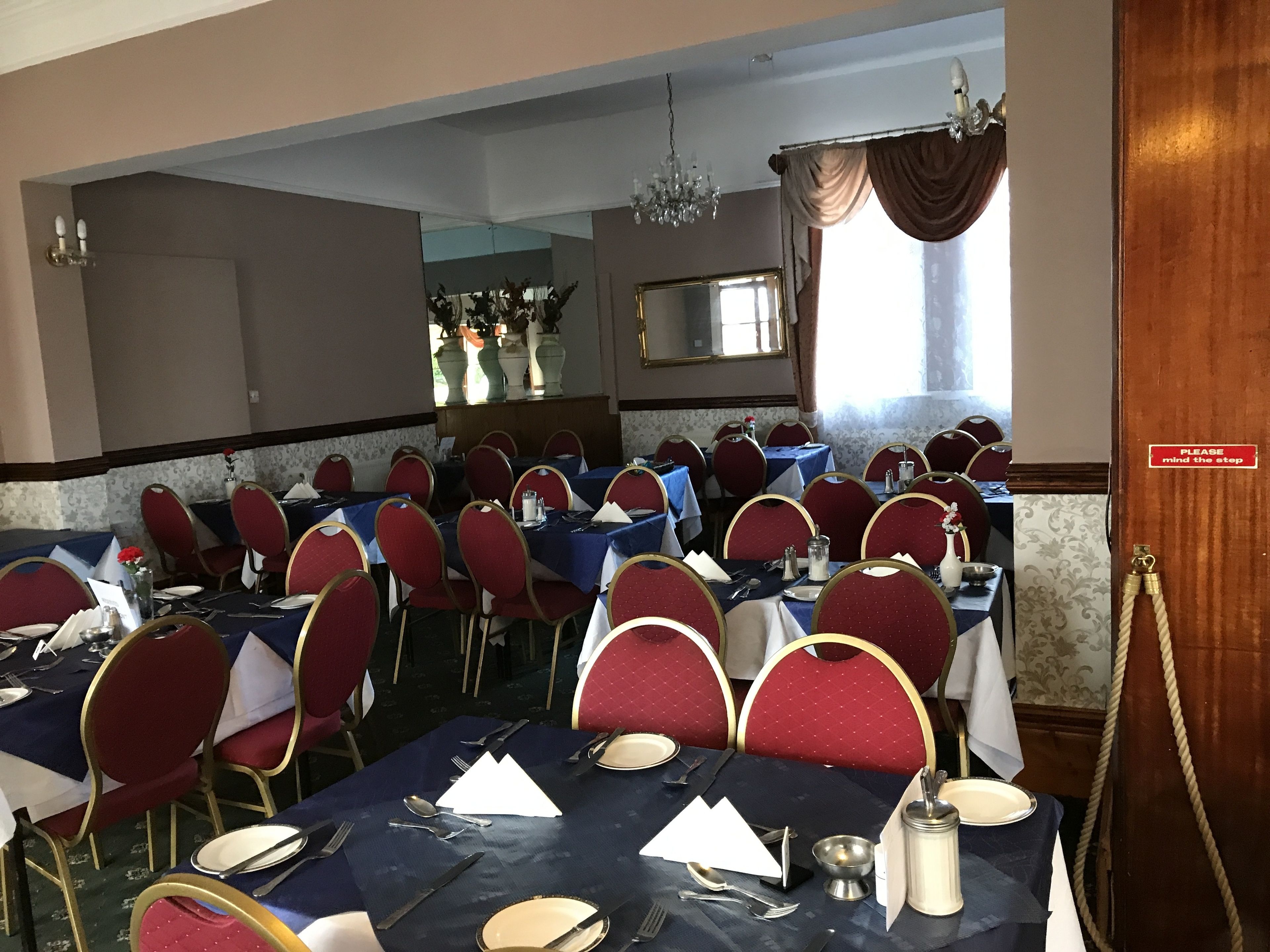 A dining area with tables set for multiple guests, red upholstered chairs, and a chandelier.
