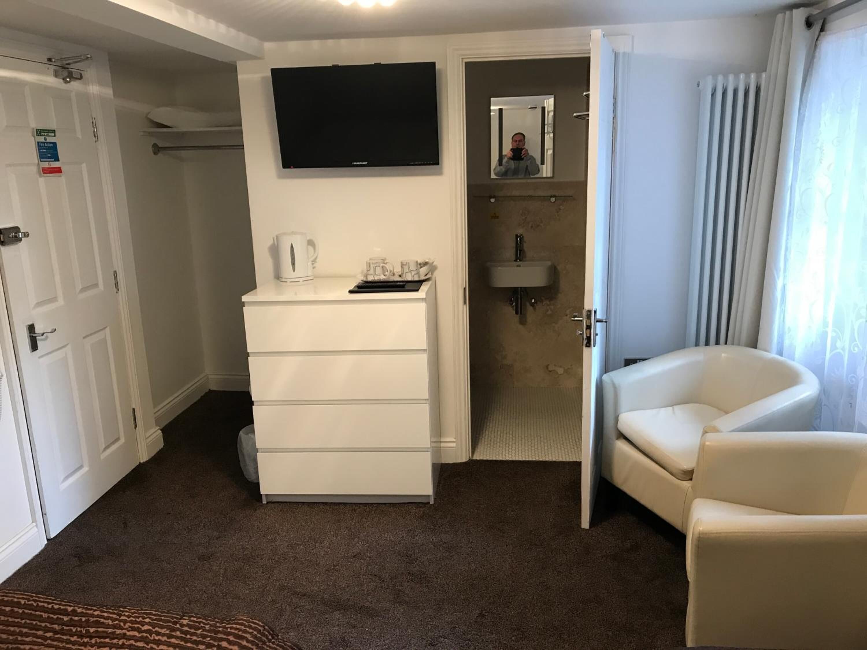 A hotel room with a flat-screen TV mounted above a white dresser, a white chair, and a bathroom visible through an open door.