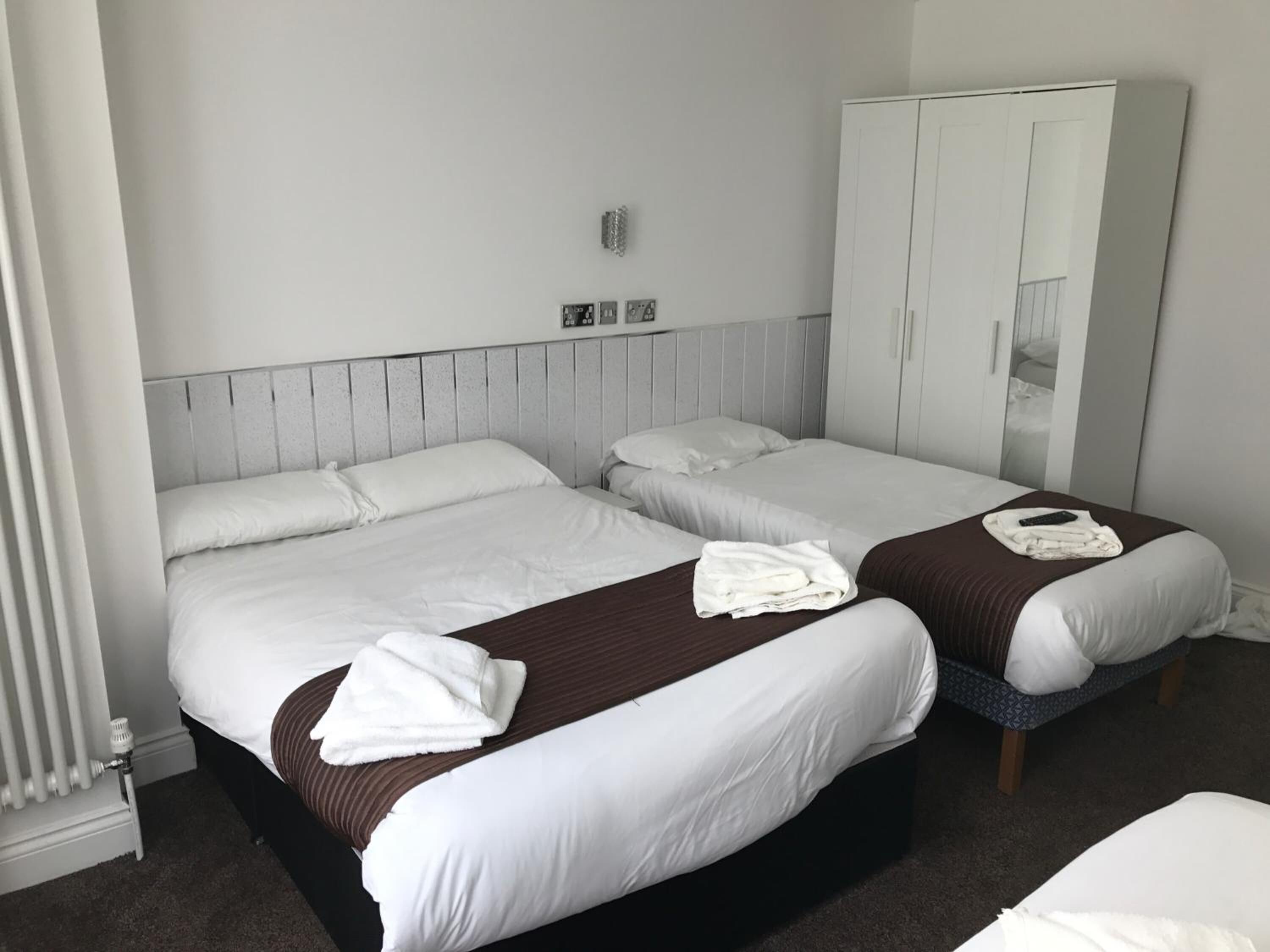 Habitación de hotel con dos camas, ropa de cama blanca, cabeceros marrones, un armario blanco y un radiador.