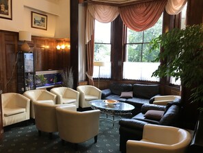 A cozy living room with leather sofas, armchairs, and a glass coffee table. A fish tank is on the left, and there are large windows with curtains. - St George Hotel (Great Yarmouth)
