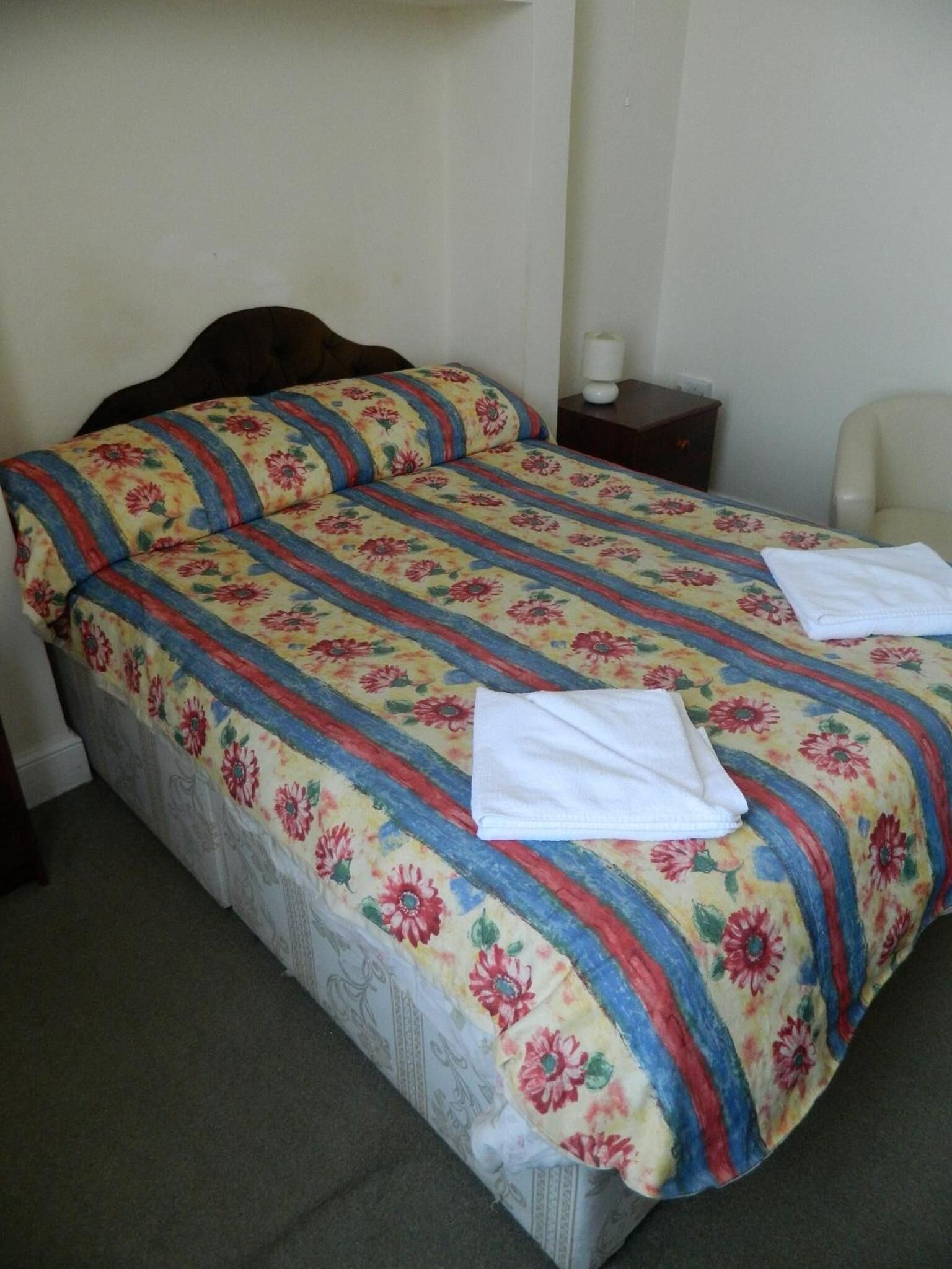 A bed with a floral-patterned bedspread, two pillows, a white towel, and a bedside table with a lamp.
