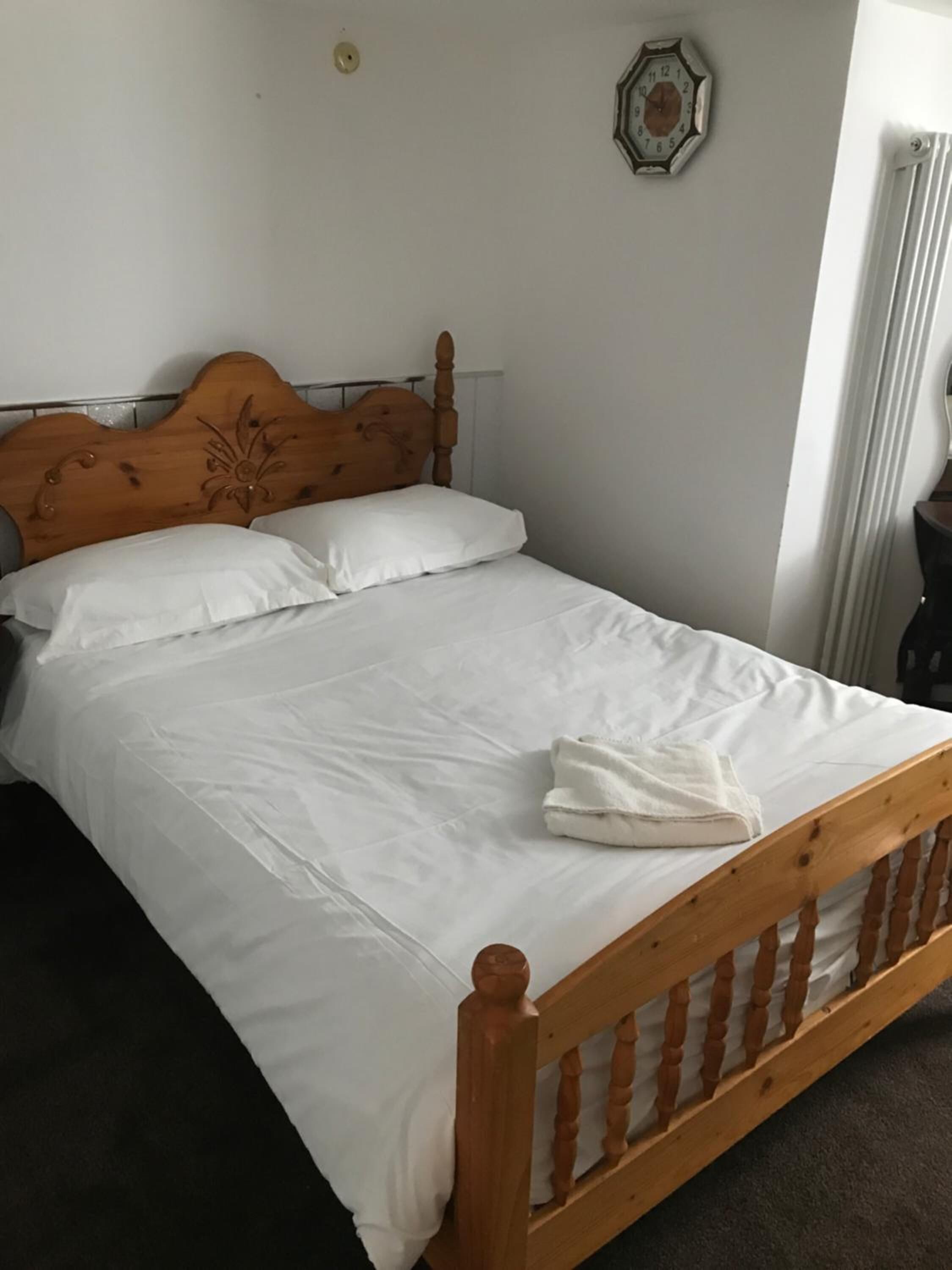 Ein ordentlich bezogenes Bett mit weißen Leinen und einem Holzkopfende, eine weiße Wanduhr und eine Heizung.