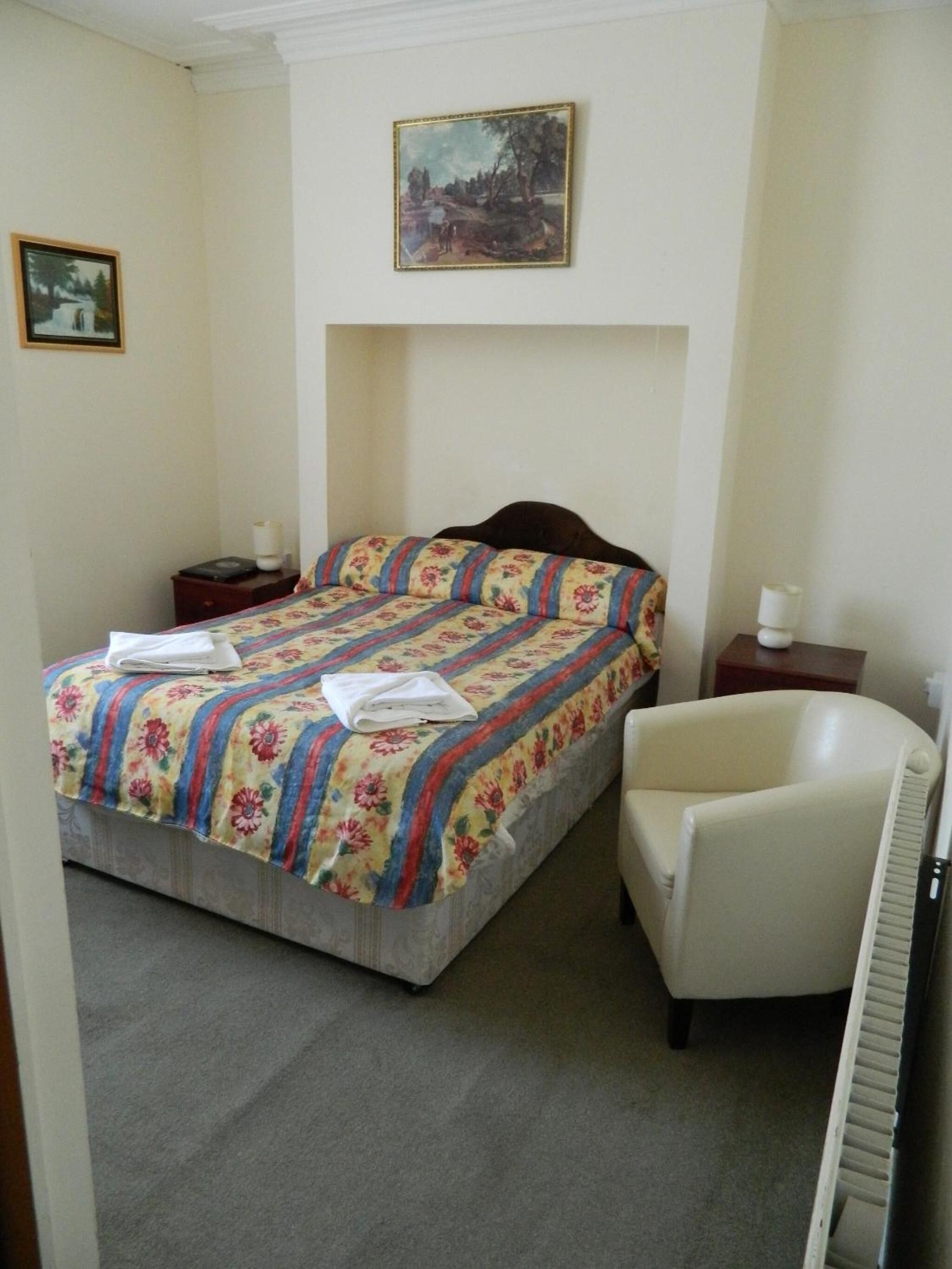 Un dormitorio con cama, una silla, una mesita de noche, una lámpara y un cuadro en la pared.