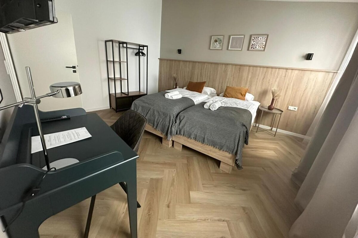 Una habitación de hotel con una cama, un escritorio con una silla, un espejo y una pequeña mesa con un florero.