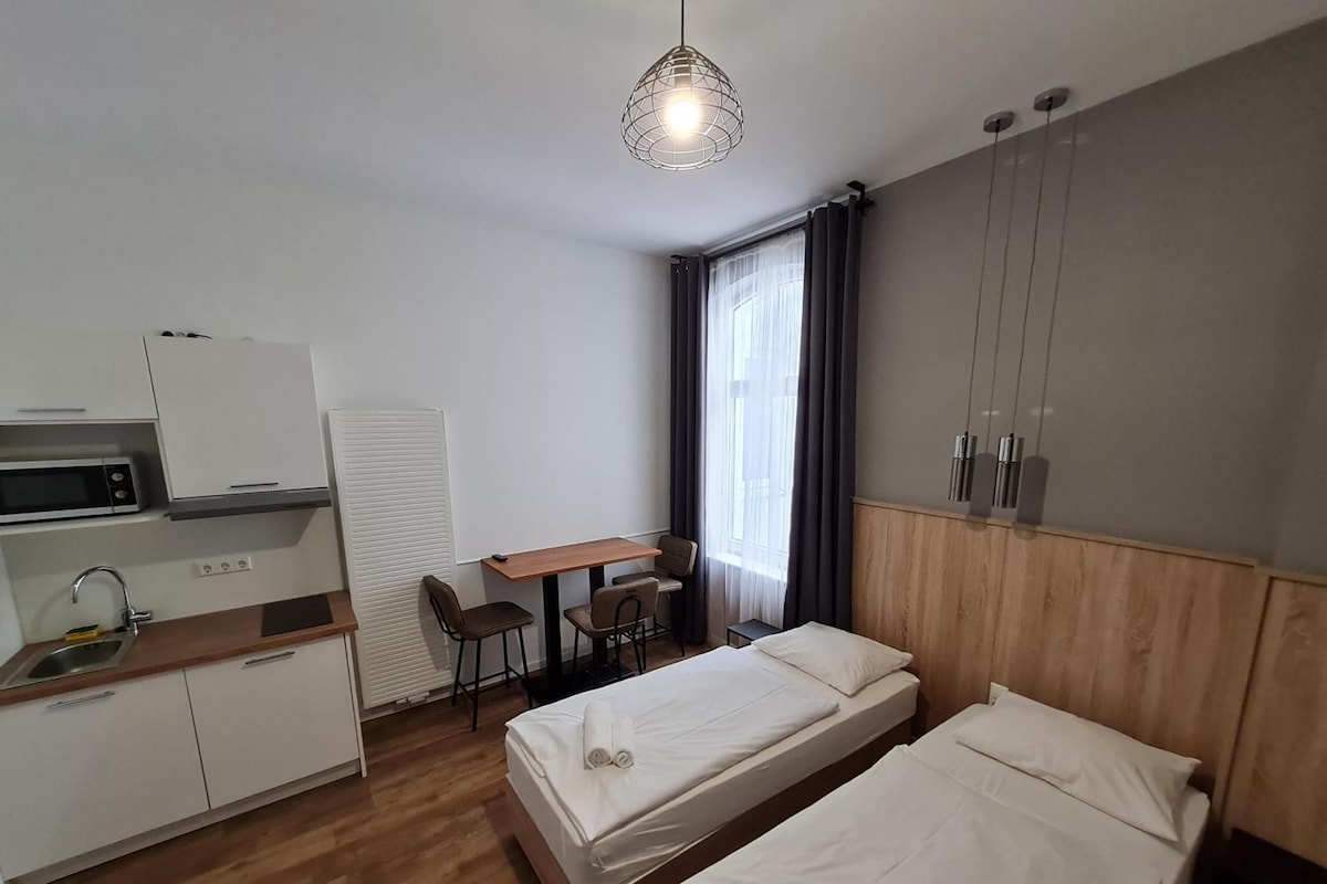 Ein Hotelzimmer mit zwei Einzelbetten, einer Küchenzeile, einem Schreibtisch mit Stühlen und einem großen Fenster mit Vorhängen.