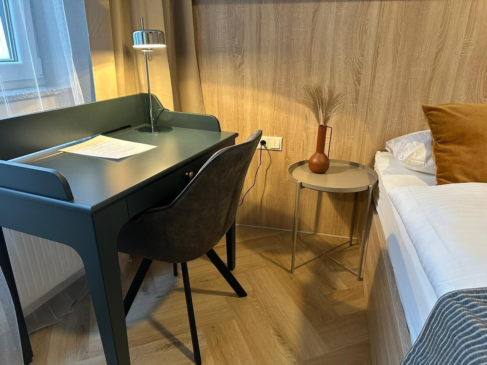Hotelkamer met een bureau, stoel, nachtkastje, lamp en een bed met kussens.