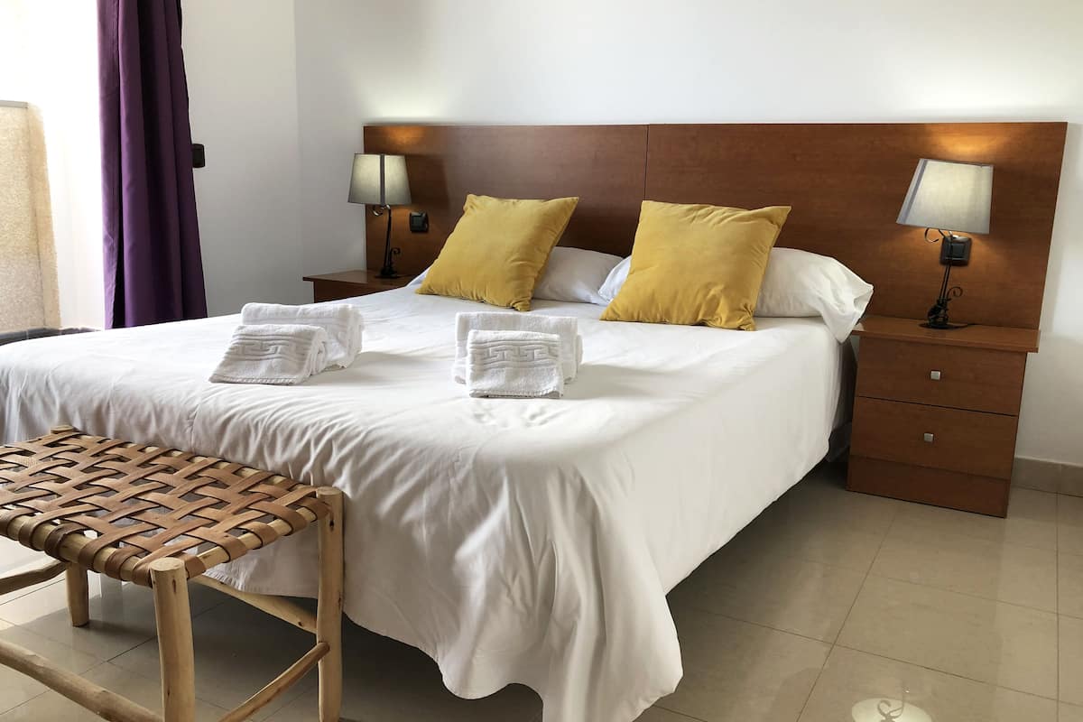 A neatly made bed with white linens and two yellow pillows, a wooden nightstand with a lamp, and a woven bench.
