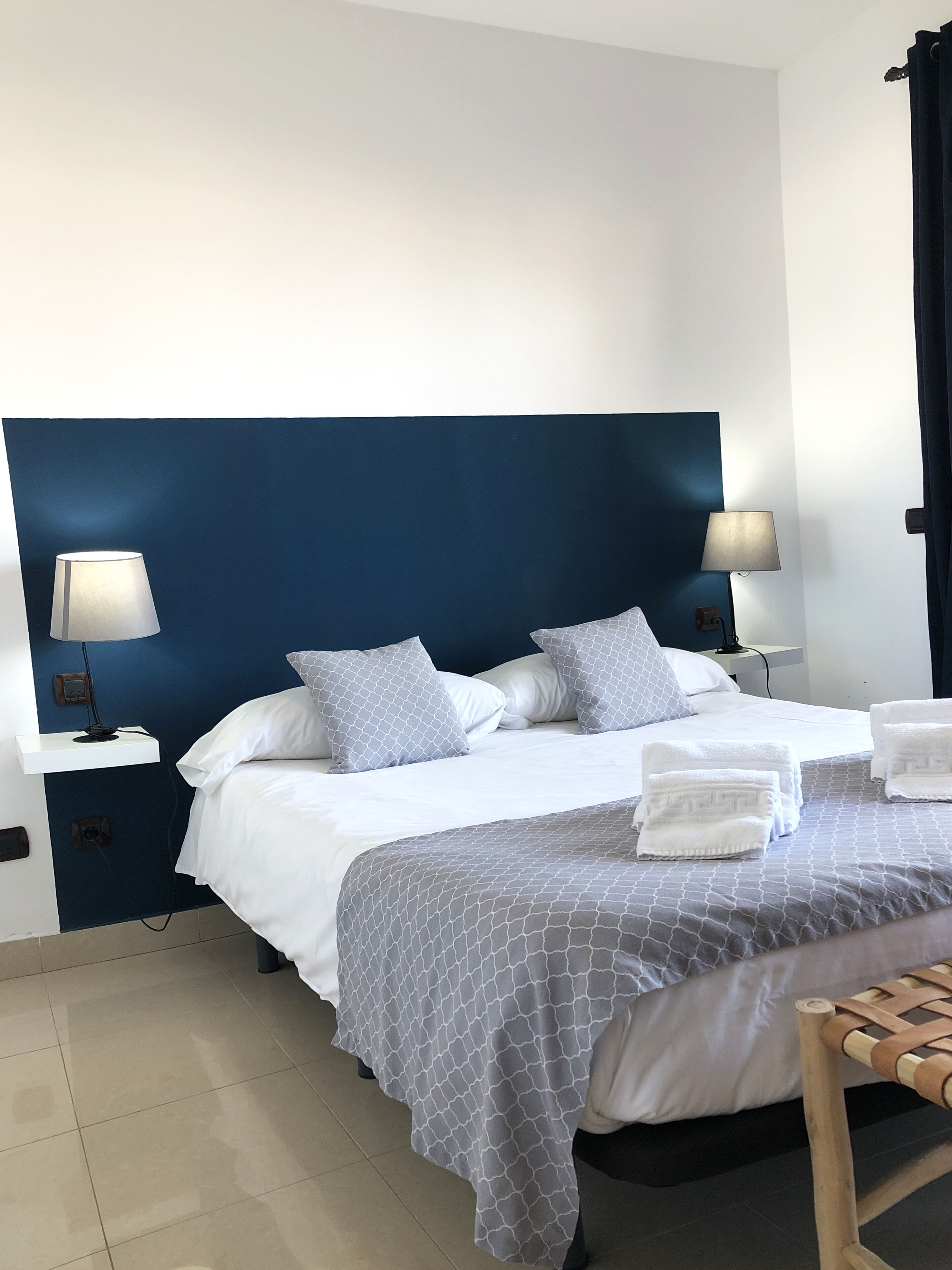 A neatly made bed with white linens and a grey patterned bedspread, flanked by two bedside tables with lamps, in a room with a dark blue accent wall.
