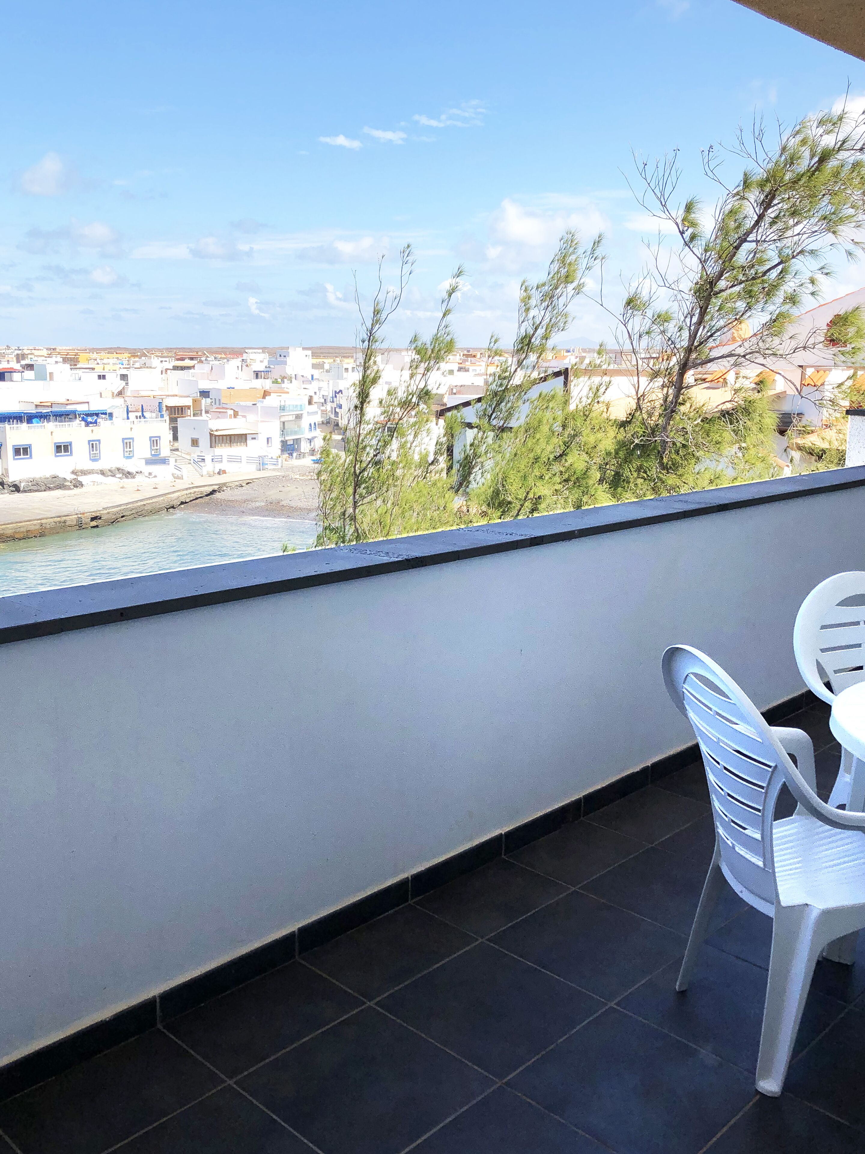 A balcony with a view of a coastal town, a table, and a chair.