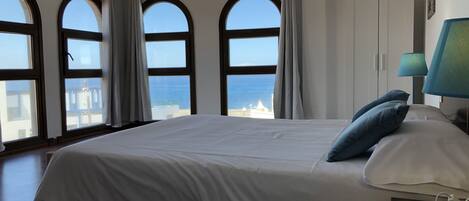 A bedroom with a large bed, a view of the sea through arched windows, and a lamp on the nightstand.
