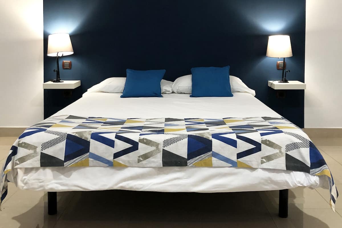A bed with a geometric-patterned bedspread, two blue pillows, and two bedside lamps on white nightstands.