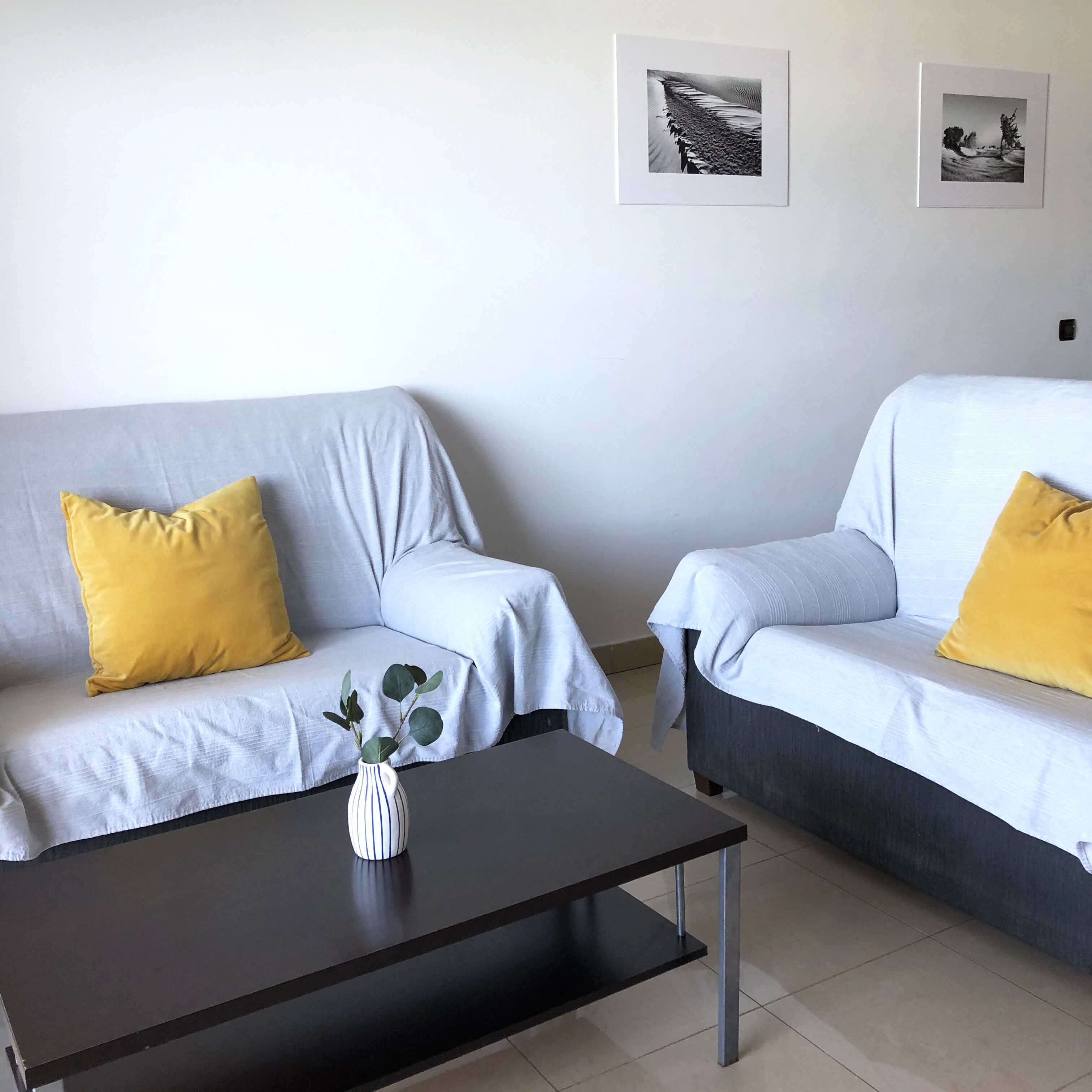A modern living room with two light grey sofas, each with a yellow pillow, a black coffee table, and a white vase with green leaves. Two framed pictures are hung on the wall.