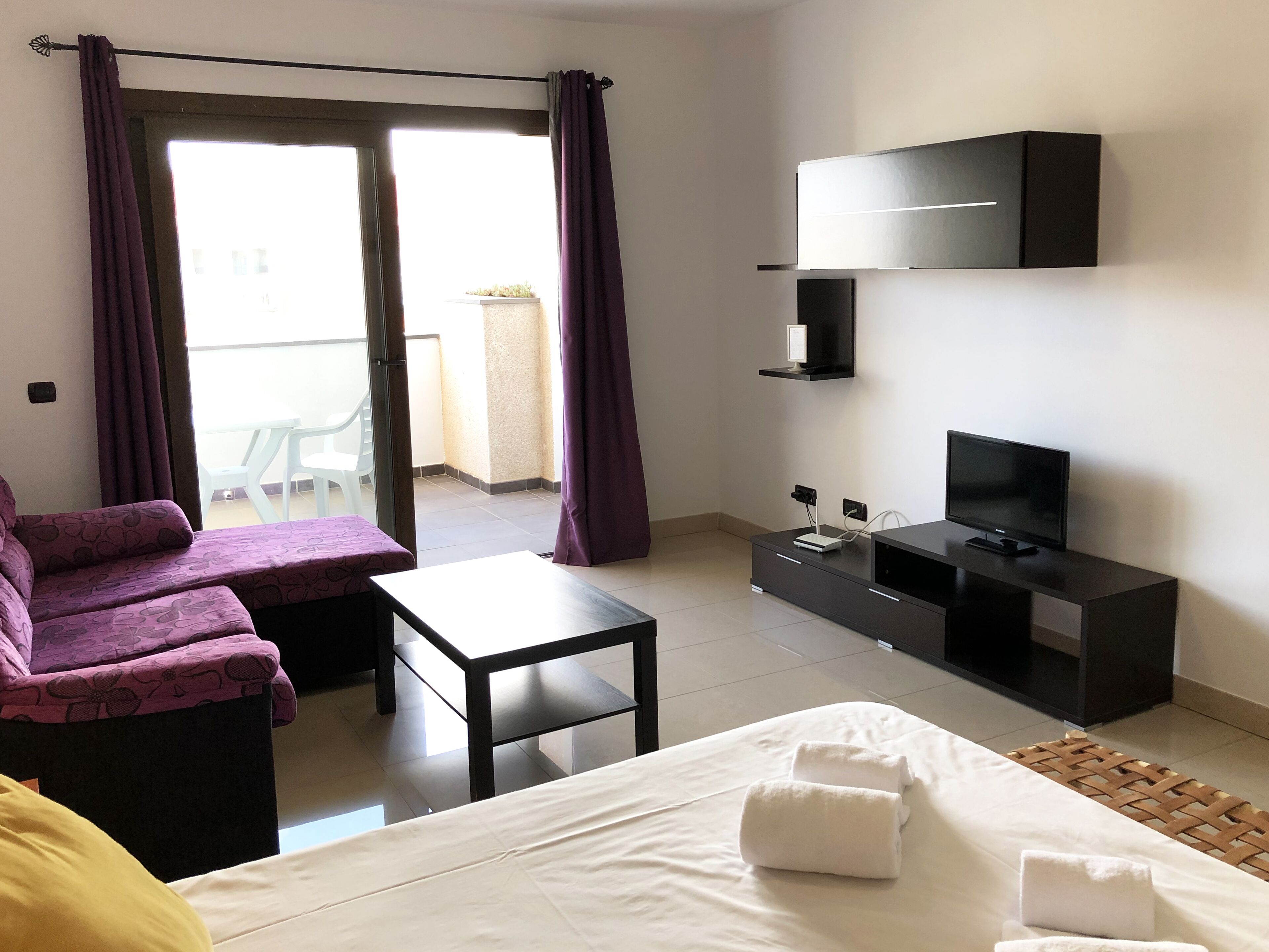A hotel room with a bed, a desk, a TV, and a balcony with purple curtains.