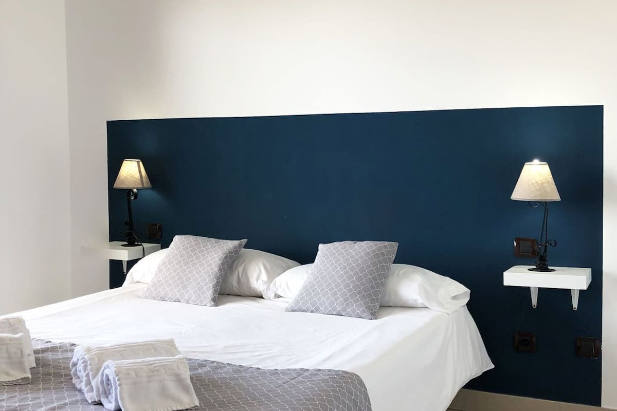 A neatly made bed with white linens and grey patterned bedding, two bedside lamps, and a small white nightstand with a lamp.
