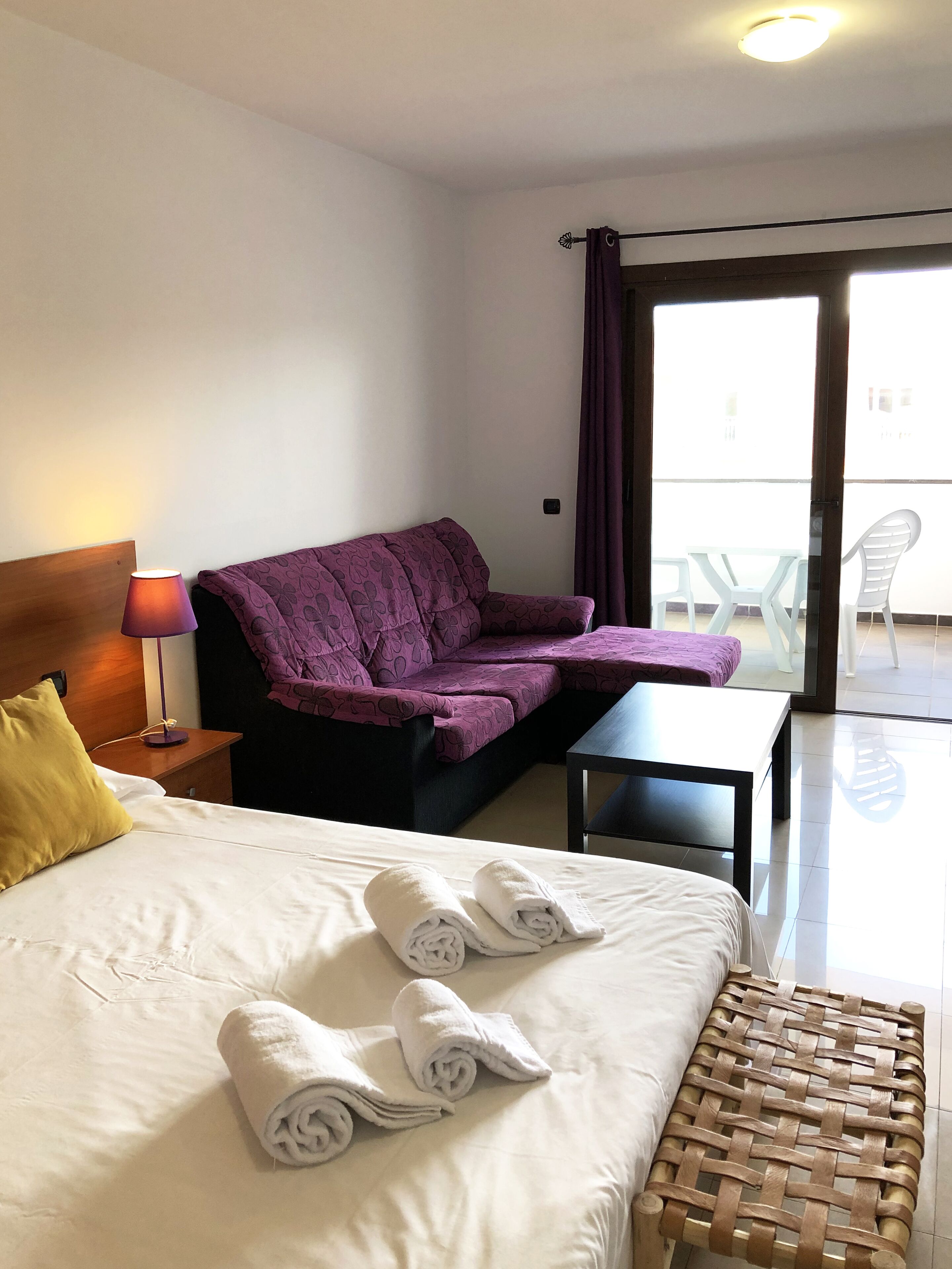 A hotel room with a bed, a sofa, a small table, and a balcony with chairs.