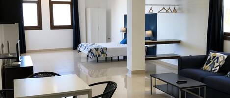 A modern living room with a sofa, coffee table, dining table, and chairs. There is a kitchen area with a counter and hanging lights.