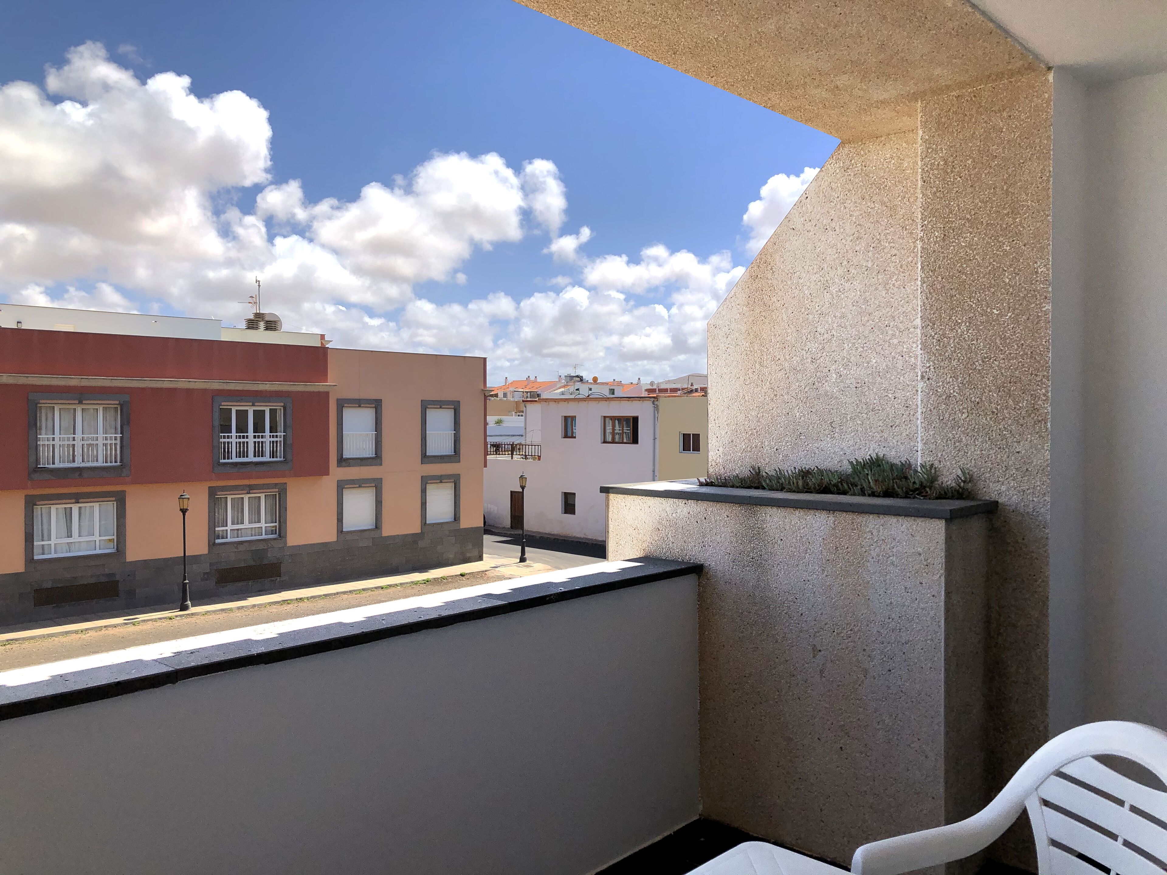 A balcony with a view of neighboring buildings, a concrete planter with plants, and a white chair.