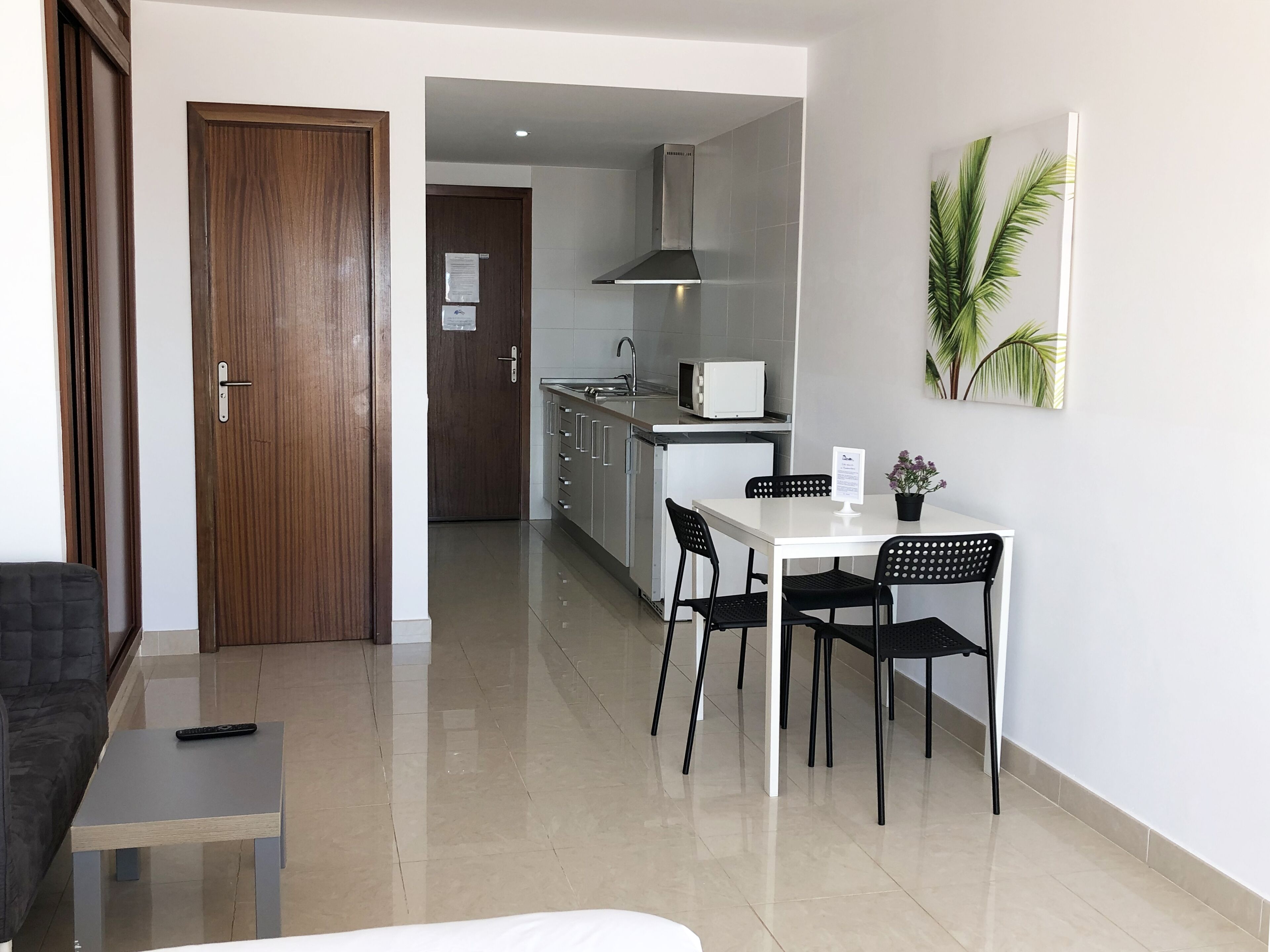 A modern kitchen with white cabinets, a microwave, and a stainless steel range hood. There is a dining area with a white table and black chairs, and a small black sofa in the living area.