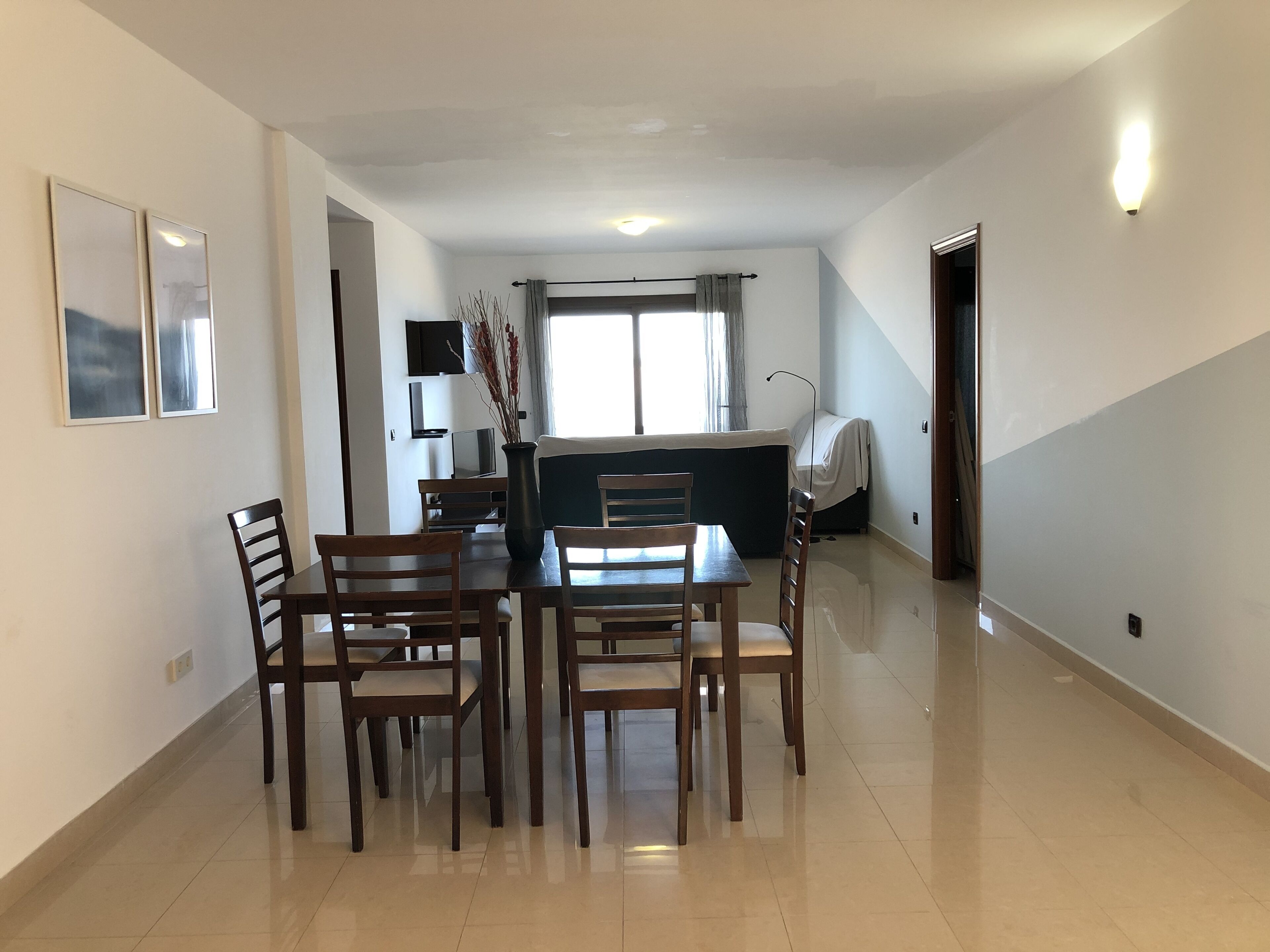 Un comedor con una mesa y sillas, un jarrón con ramas, un televisor y una ventana con cortinas.