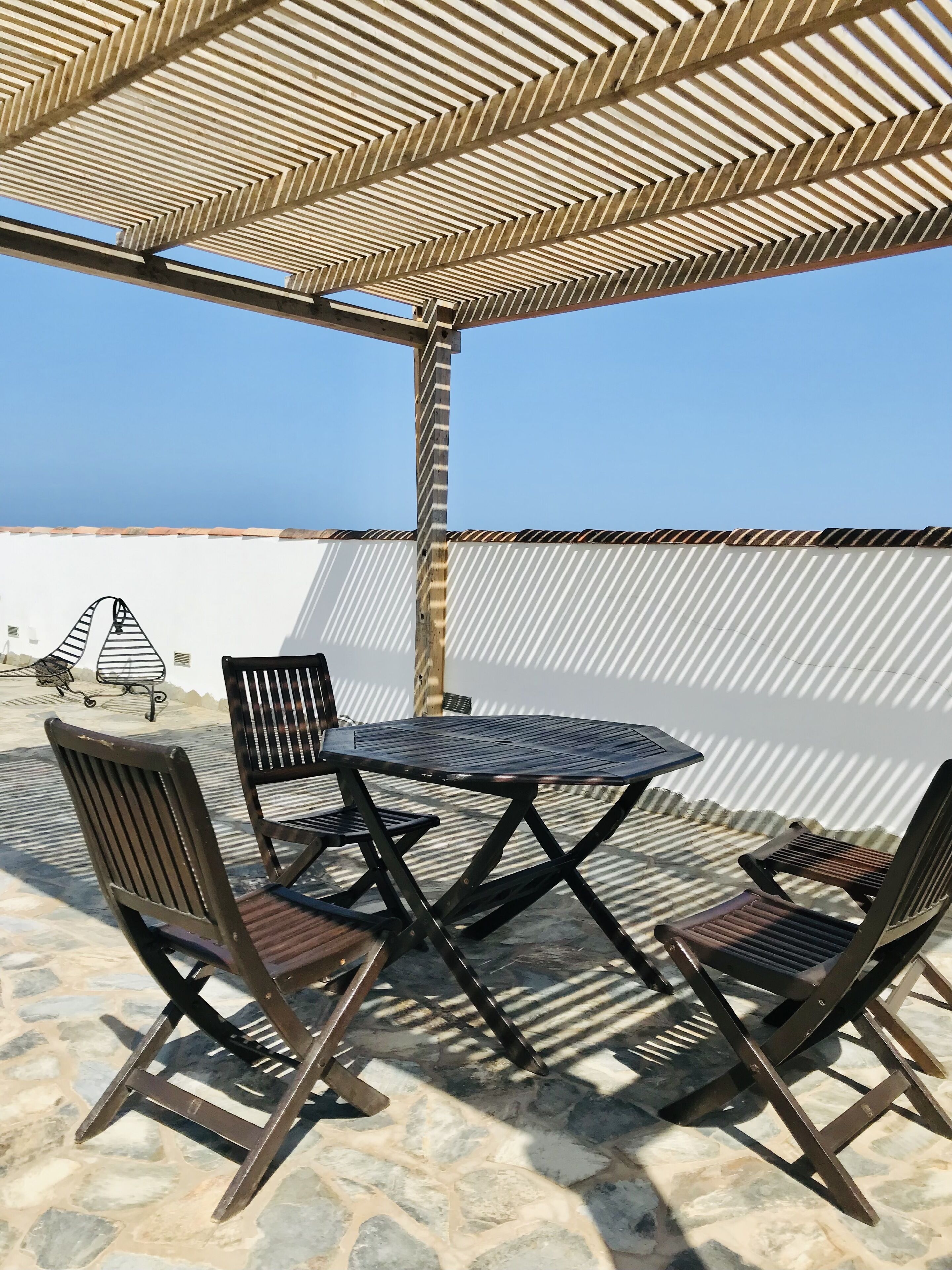 Un pati exterior amb una taula i caires, una pérgola a sobre i vista al mar.
