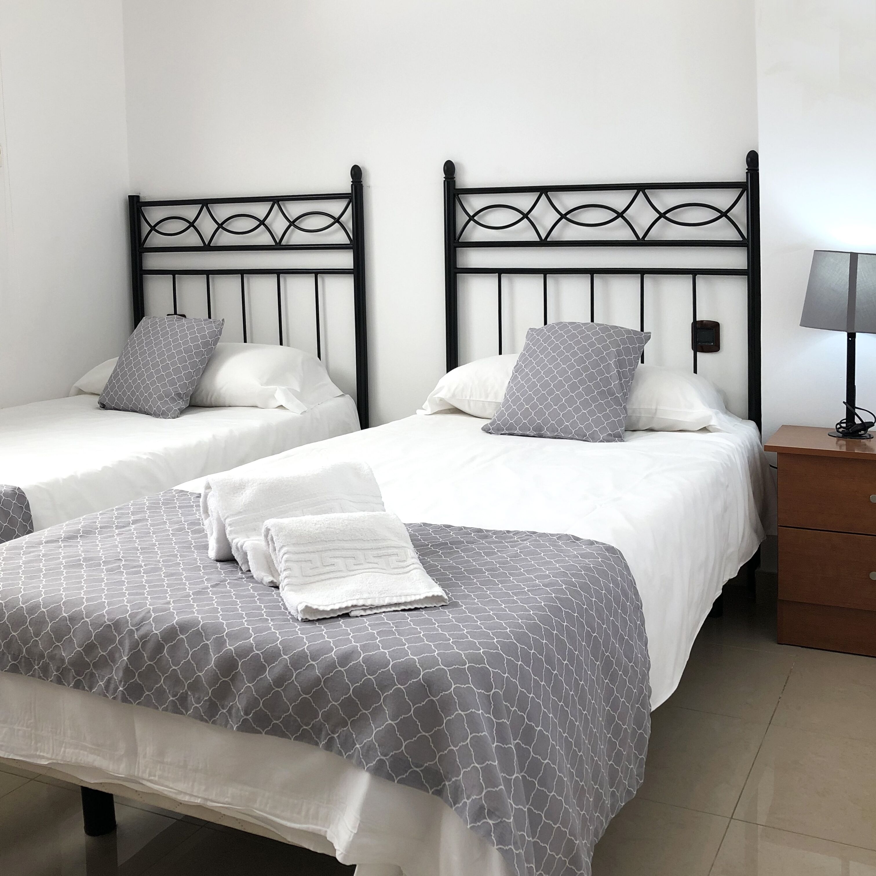 Two single beds with black metal frames and white bedding, a bedside table with a lamp, and a wooden nightstand.