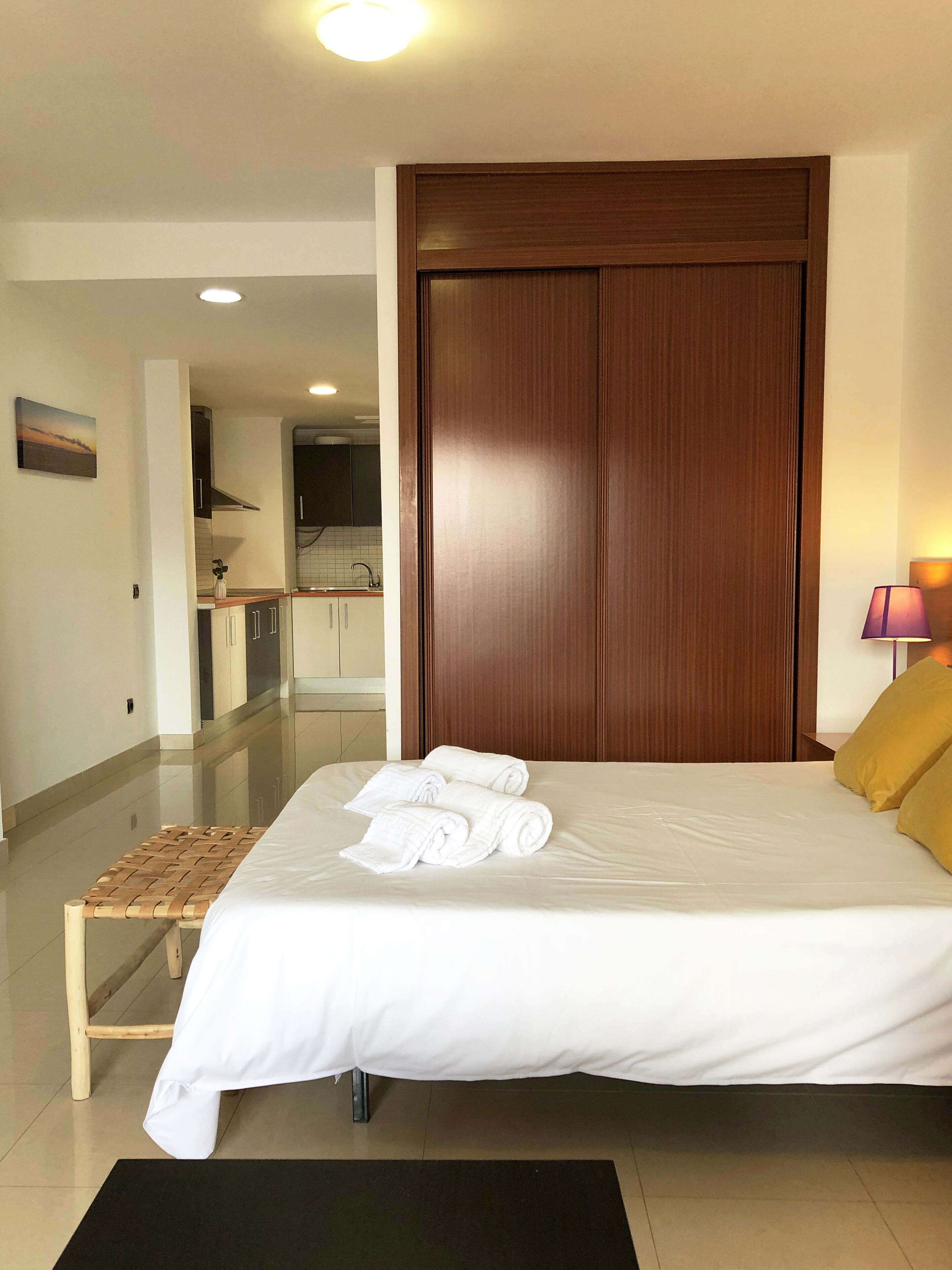 Habitación de hotel con una cama, un banco de madera, un armario de madera, una mesita de noche con lámpara y vista a la cocina.