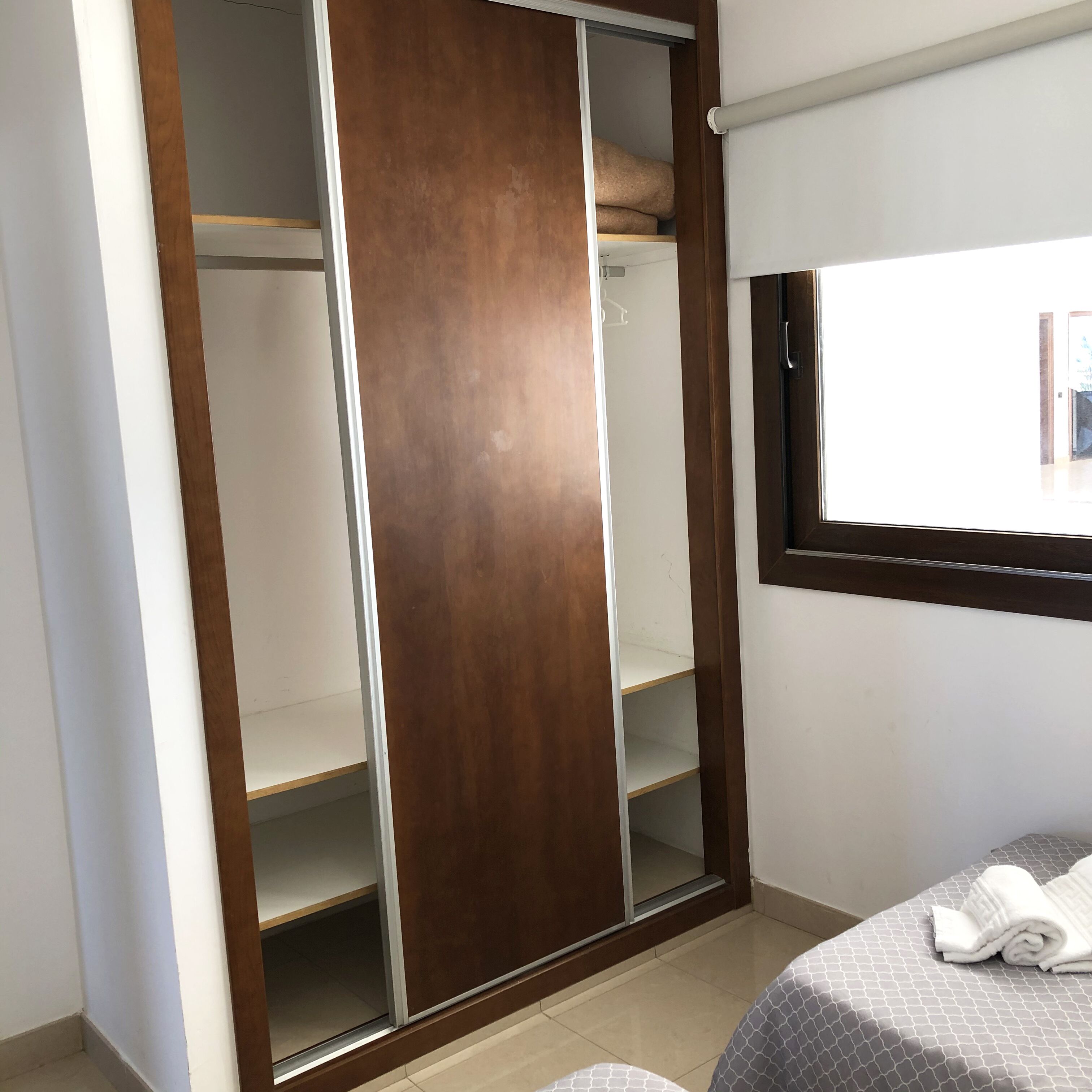 A bedroom with a wardrobe, a bed with a patterned bedspread, and a window with blinds.