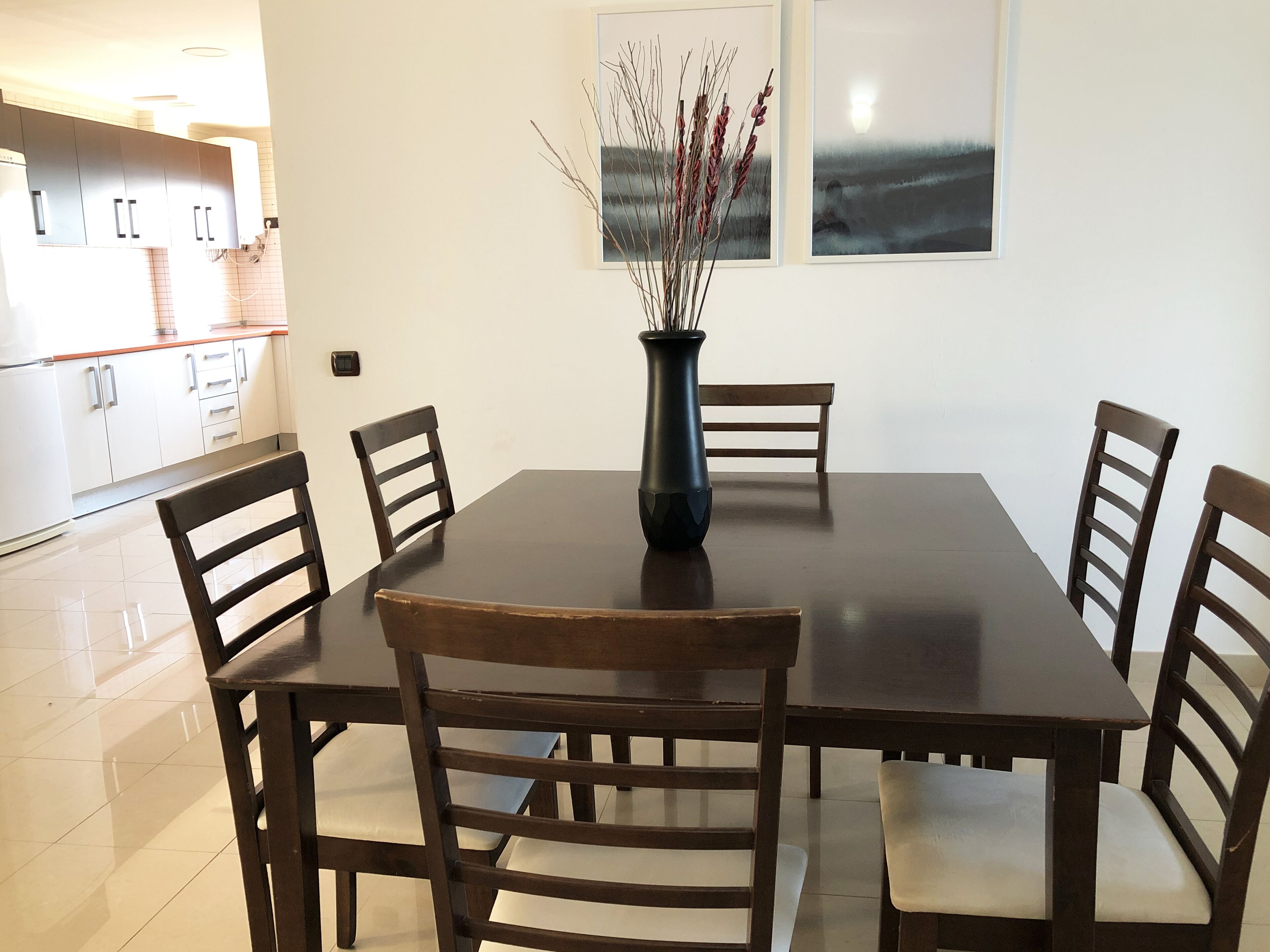 A dining area with a dark wooden table and chairs, a vase with dried plants, and two framed pictures on the wall.