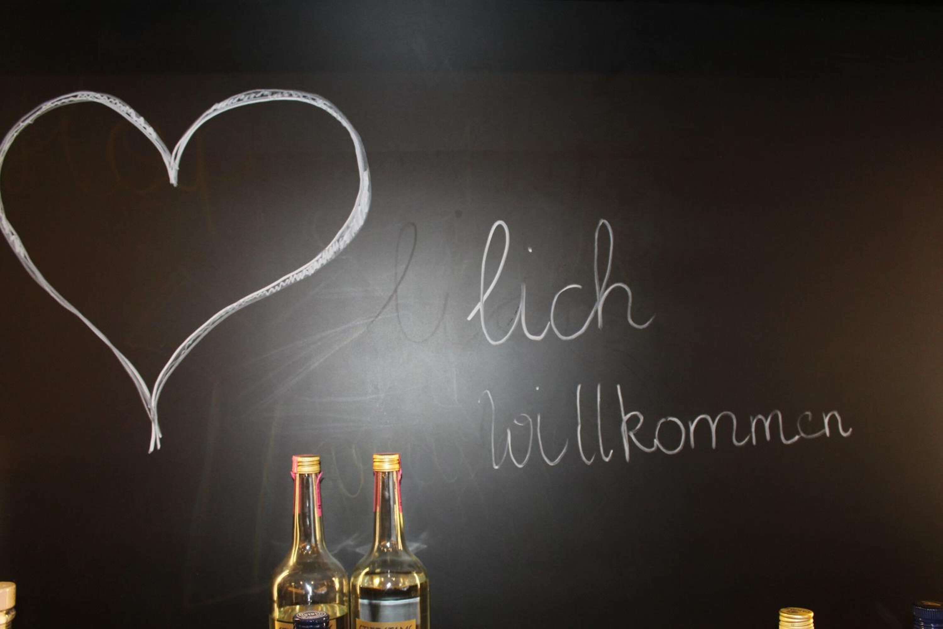 Una pizarra con un corazón y las palabras 'Ich liebe dich' y 'Willkommen', con dos botellas de vino delante.