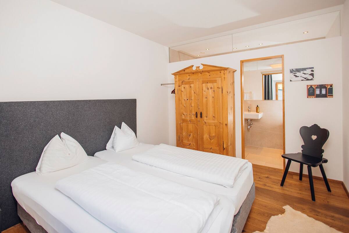 A bedroom with a large bed, a wooden wardrobe, a small chair, and a bathroom visible through an open door.