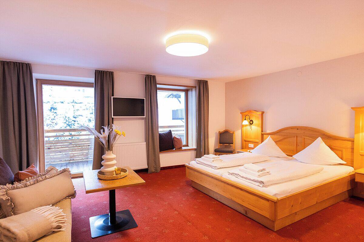 A hotel room with a bed, a TV, a window with curtains, a table with a vase, and a lamp.
