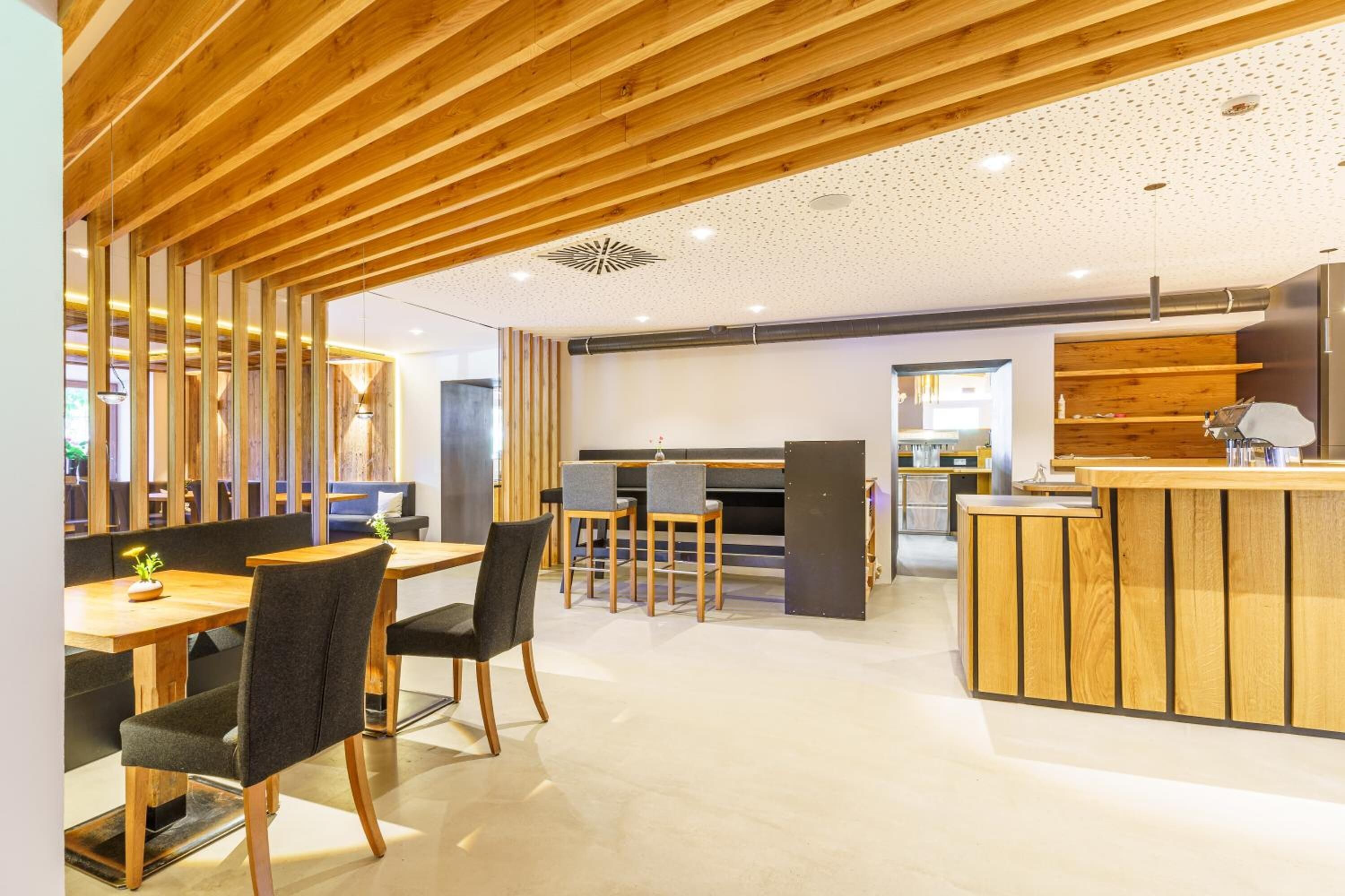 Un interior moderno con vigas de madera en el techo, un área de comedor central con sillas y una mesa, y una cocina con mostrador y electrodomésticos.
