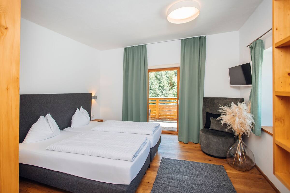 A bedroom with a bed, a chair, a TV, and a window with curtains.