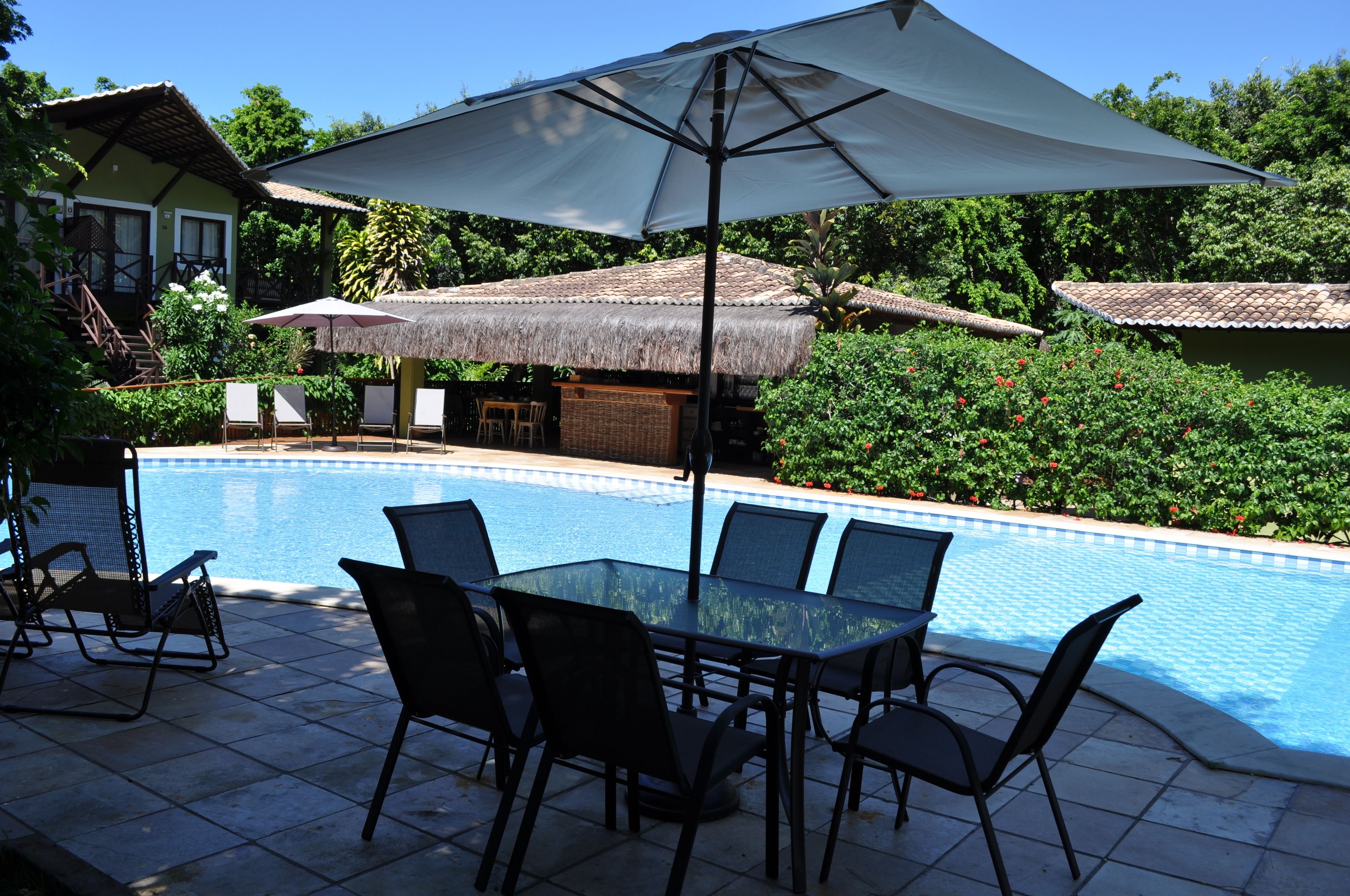 A pool area with a table and chairs, a large umbrella, and a thatched-roof structure.