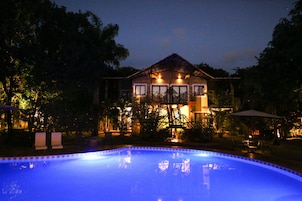 A poolside view of a hotel with a swimming pool, outdoor seating, and a well-lit building with a staircase leading to the entrance.