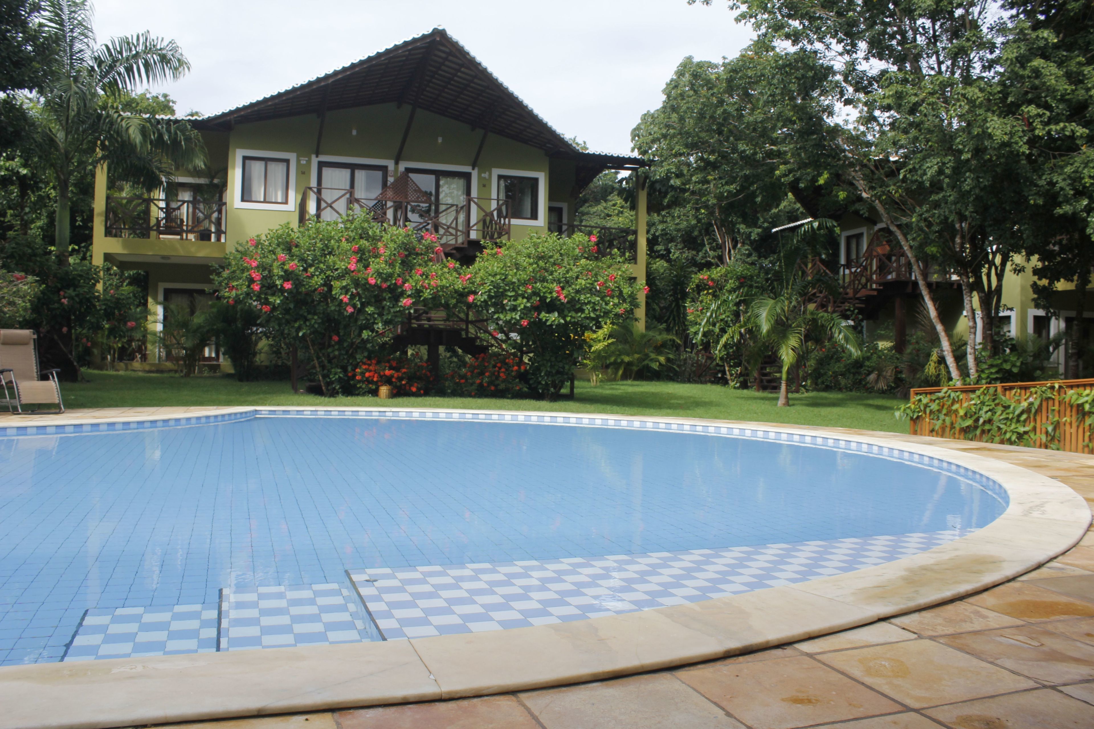 Una piscina amb un dibuix de quadres, envoltada d'un jardí amb diferents plantes i una casa amb un balcó.