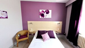 A hotel room with a bed, a chair, a bedside table, and a wall with a floral artwork. - Hôtel la Croix Couverte (Mayenne)
