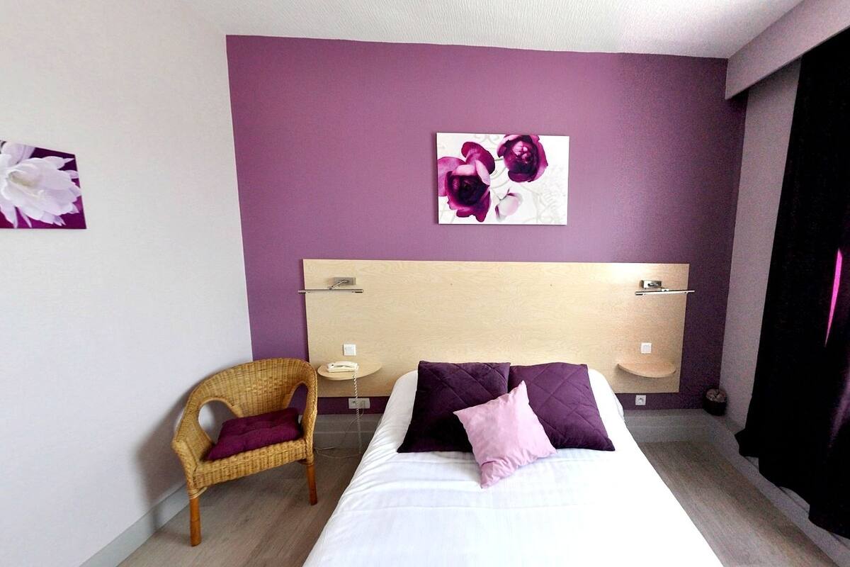 A hotel room with a bed, a chair, a bedside table, and a wall with a floral artwork.