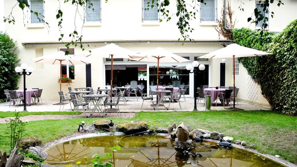 An outdoor patio area with tables and chairs, umbrellas, and a small pond with rocks and a statue. - Hôtel la Croix Couverte (Mayenne)