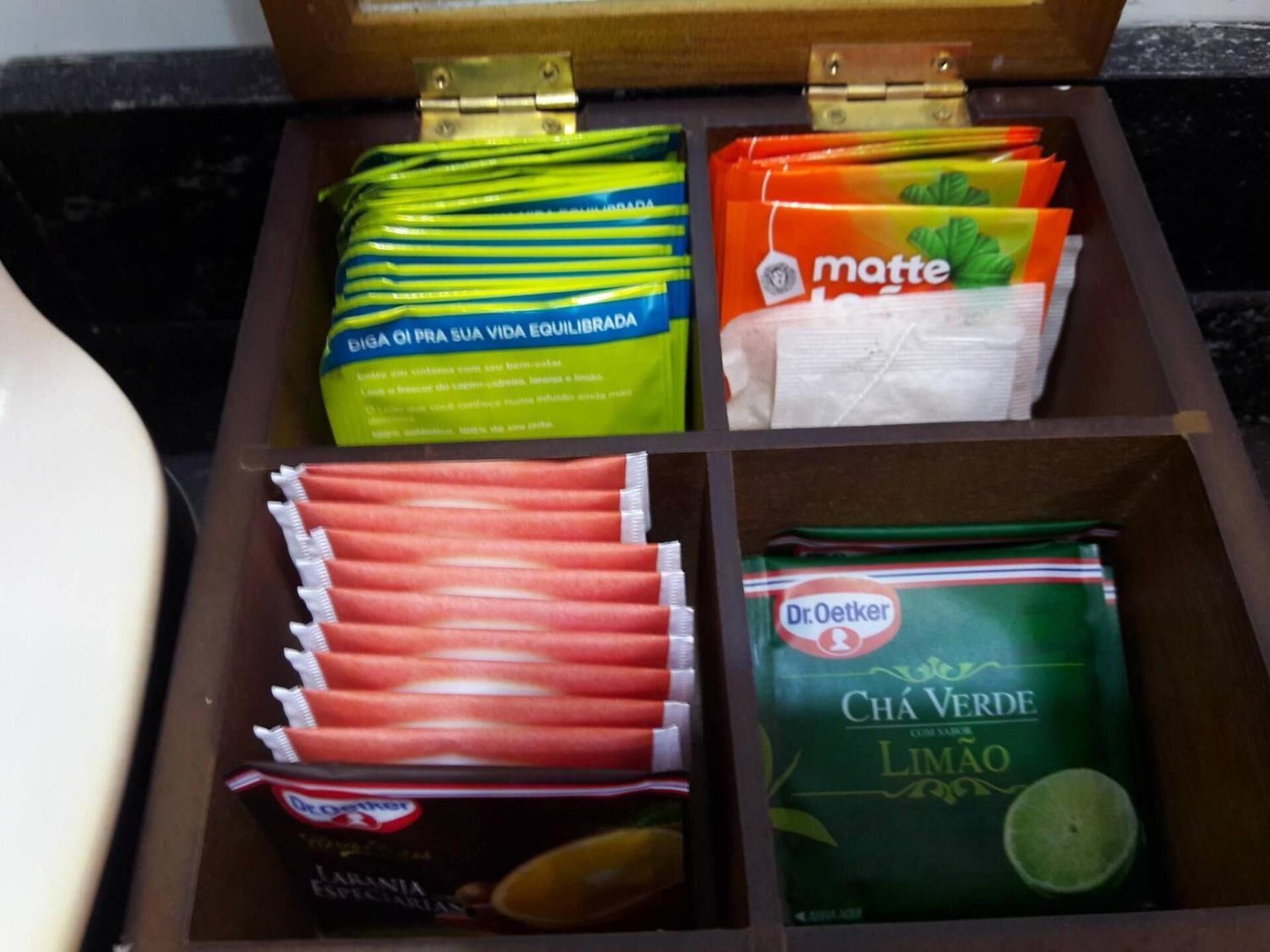 A wooden box containing various food items including packets of Biga OI, matte, Dr. Oetker Chá Verde Limão, and other snacks.