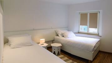 A bedroom with two beds, a nightstand, a lamp, a window with blinds, and a wooden floor.