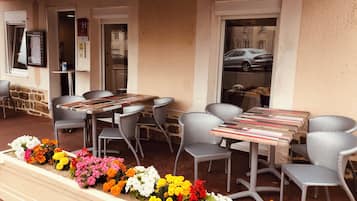 Un área de estar al aire libre con sillas y mesas de mimbre, un parterre con diversas flores y un edificio con un letrero que dice 'STAURANT'.