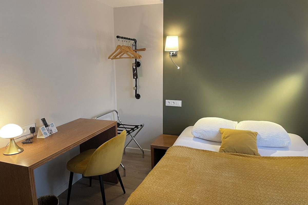 A hotel room with a wooden desk, a yellow chair, a bed with a brown blanket, and a wall-mounted lamp.