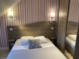 Quarto de hotel com cabeceira de madeira, duas luminárias de apoio, cama com lençóis brancos e almofadas estampadas, e piso de madeira.