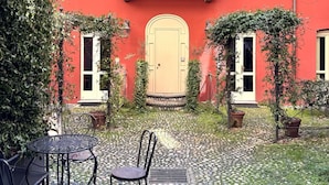 A courtyard with a red building, a door, and two windows. There are potted plants and a table with chairs. - Residence Porta Palace - Welkome Apartments (Turin)
